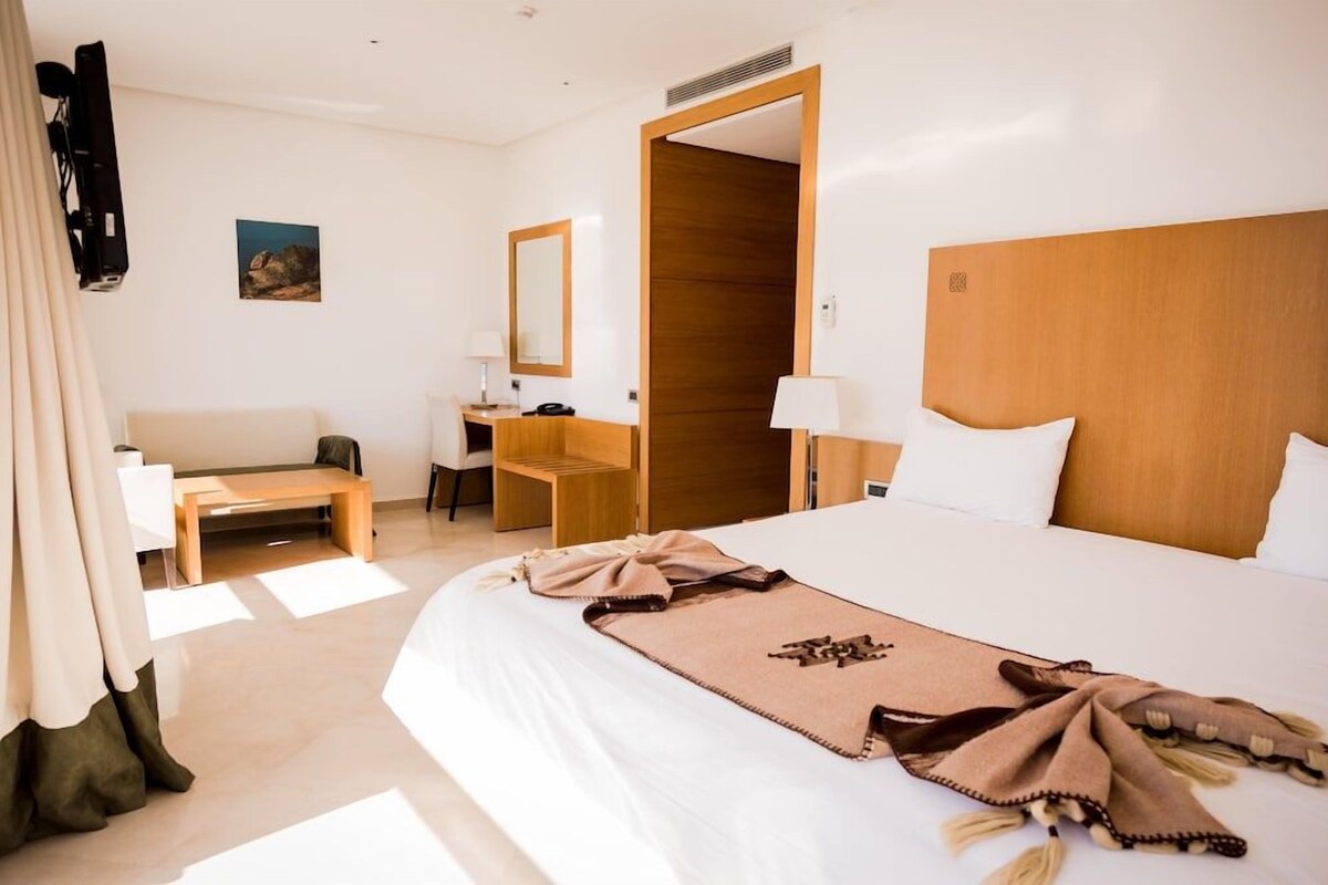 A hotel room with a large bed, a desk, a chair, a sofa, and a wooden wardrobe.