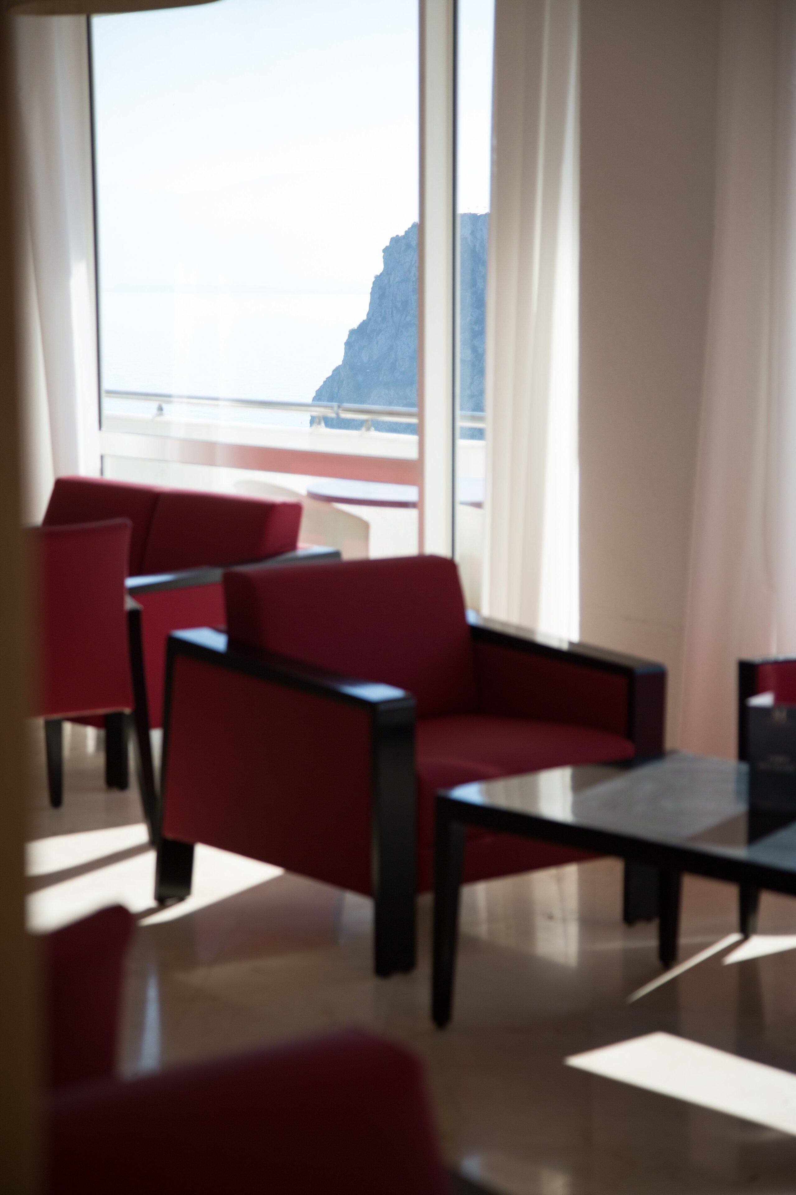 Una habitación con sillas rojas y una mesa, ventanas amplias con vista al mar y un acantilado.