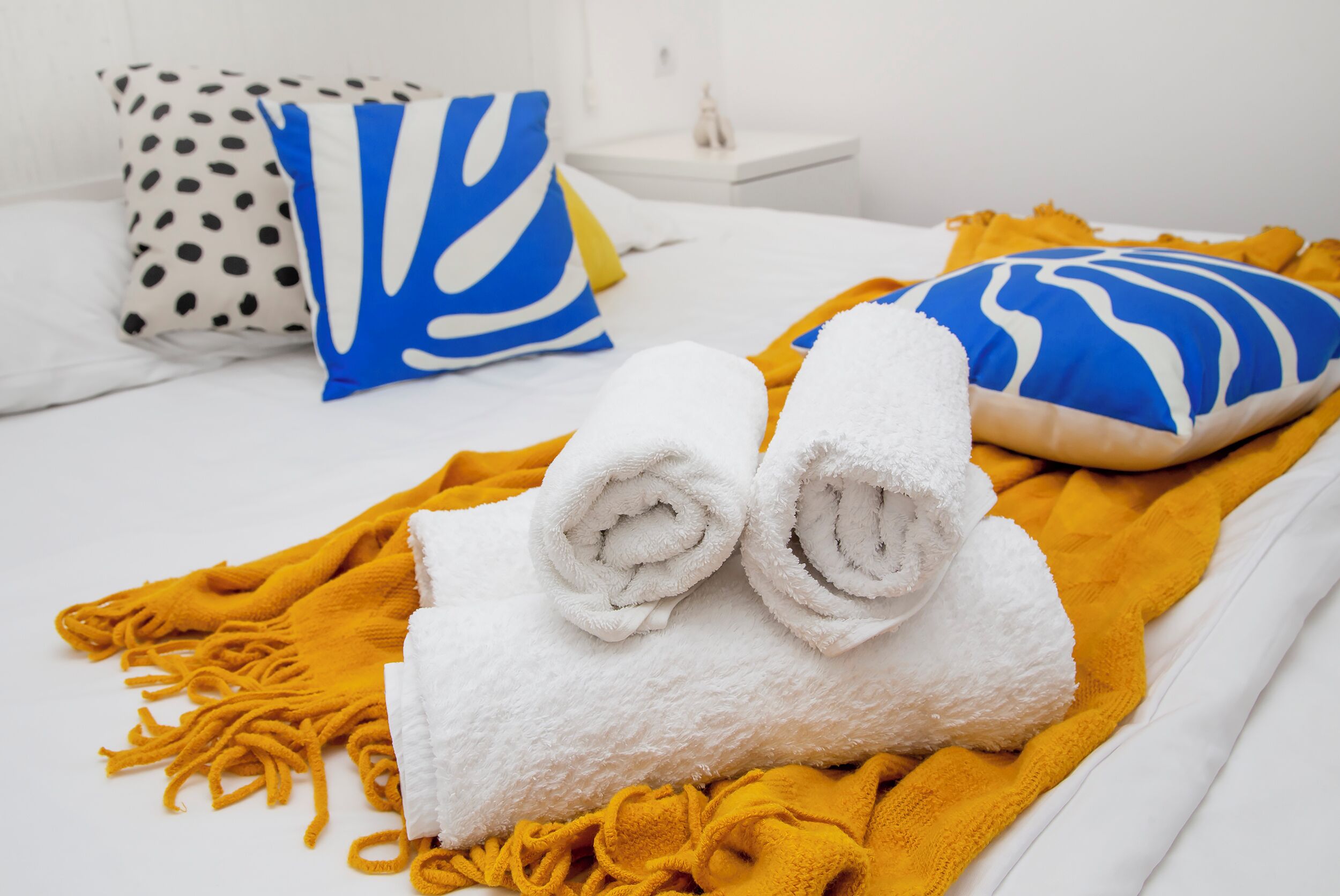 A neatly made bed with rolled white towels, a yellow throw blanket, and two decorative pillows with blue and white patterns.