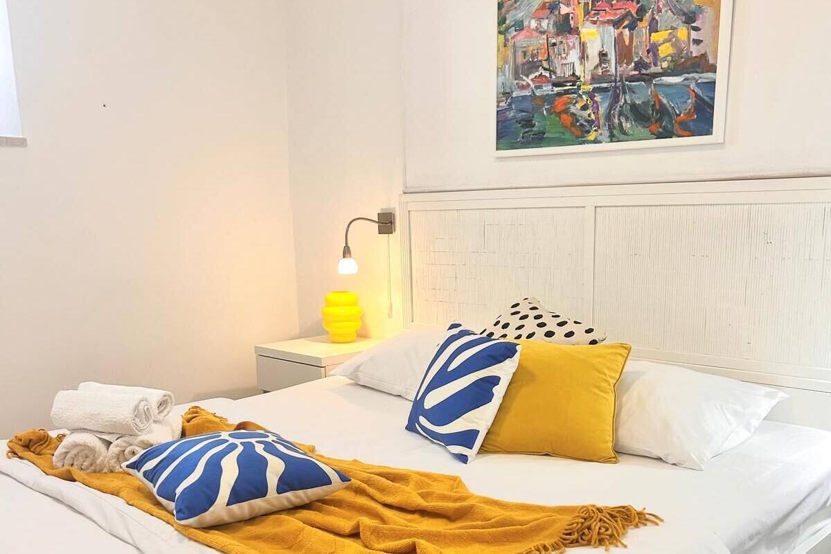 A neatly made bed with white linens, a mustard throw blanket, and patterned pillows. A painting hangs above the bed, and a bedside lamp is visible.