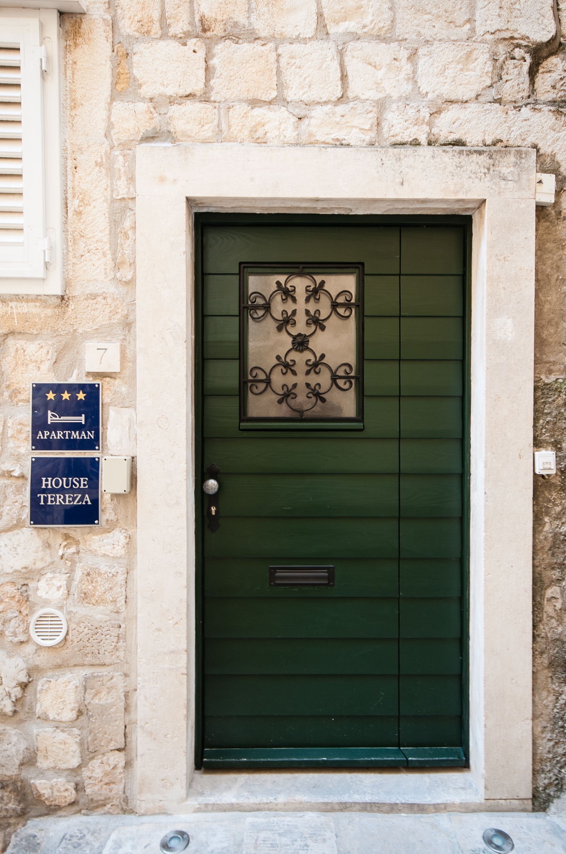 Una porta verde con un pannello decorativo, un cartello con il numero 7 e due insegne che indicano “APARTMAN” e “HOUSE TEREZA”.
