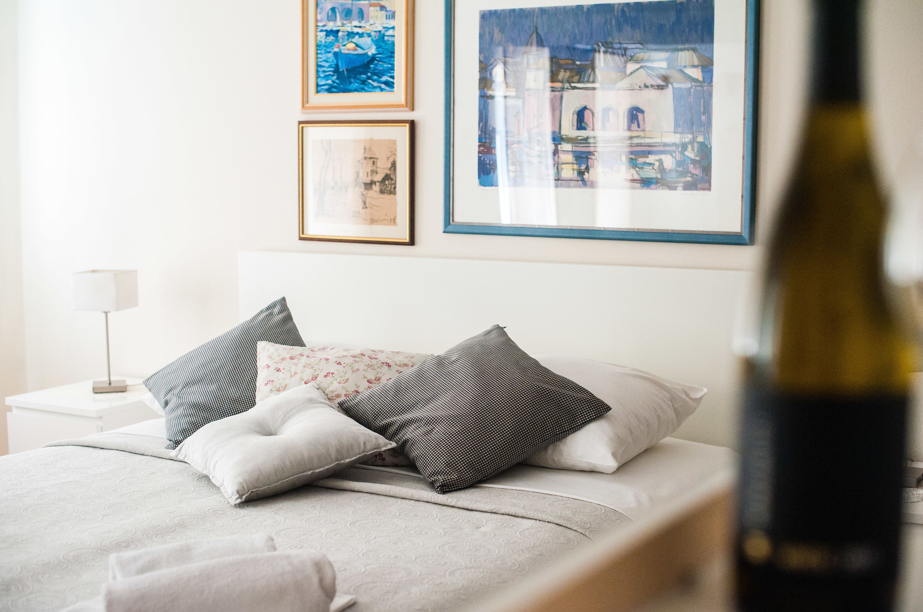 Una cama bien hecha con almohadas, una mesita de noche con lámpara y cuadros en la pared.