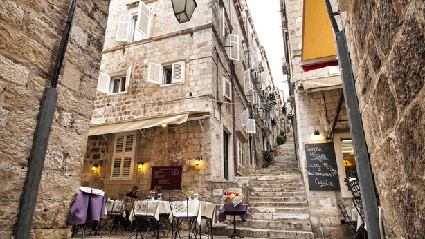 Una calle estrecha con edificios de piedra, mobiliario exterior con mesas y sillas, y una escalera que sube a un nivel superior.