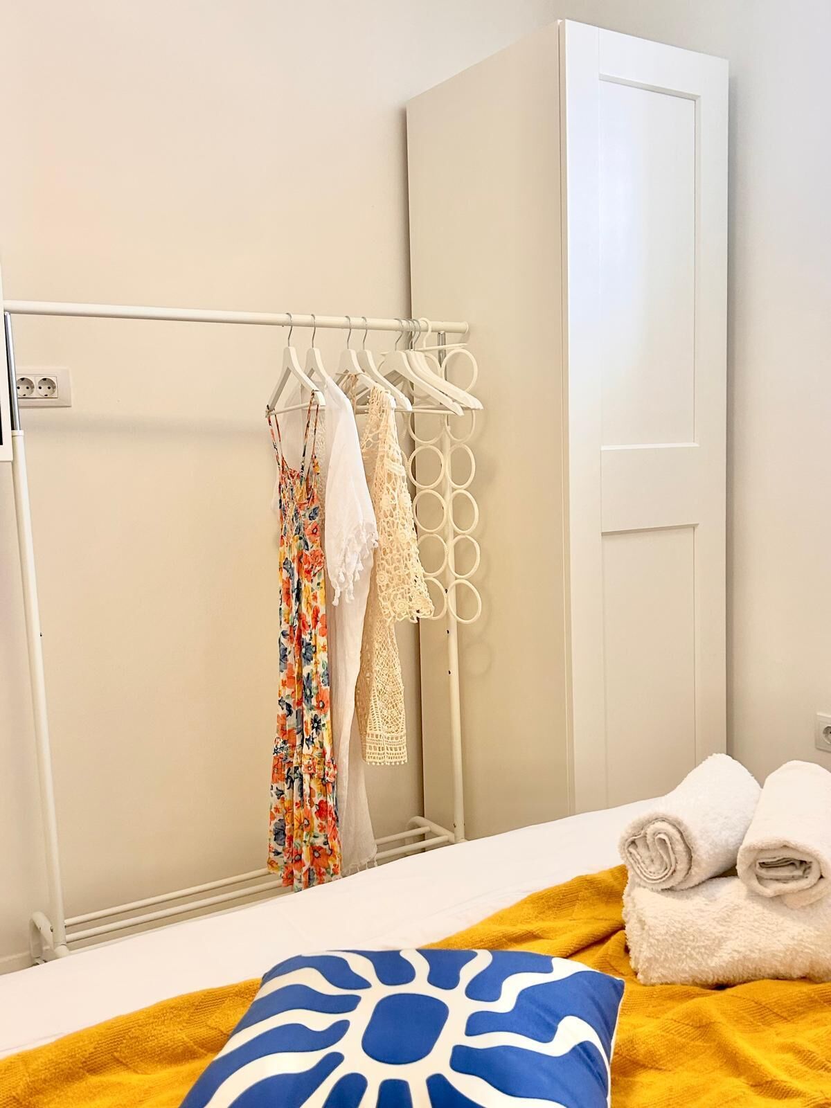 A hotel room with a bed, a white wardrobe, a clothing rack with various garments, and a rolled-up towel on the bed.