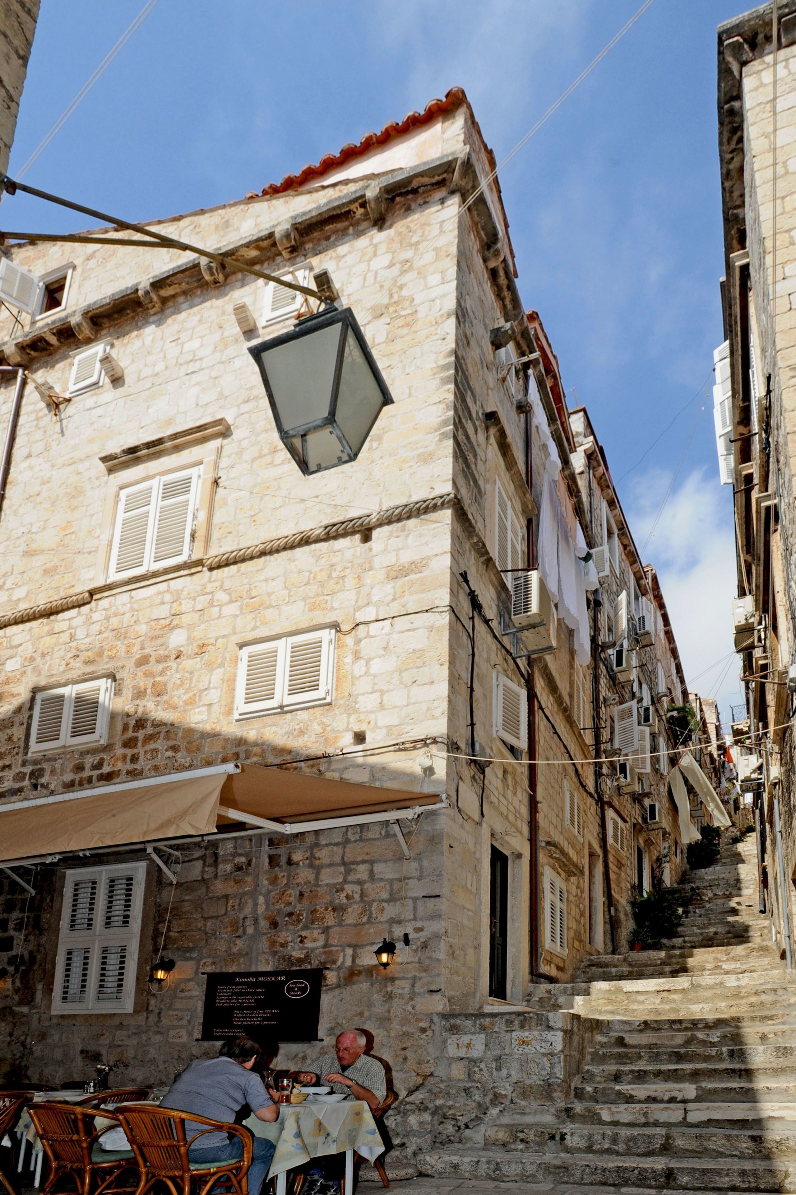 Una estrecha calle con edificios de piedra, mesas al aire libre y una escalera que sube.