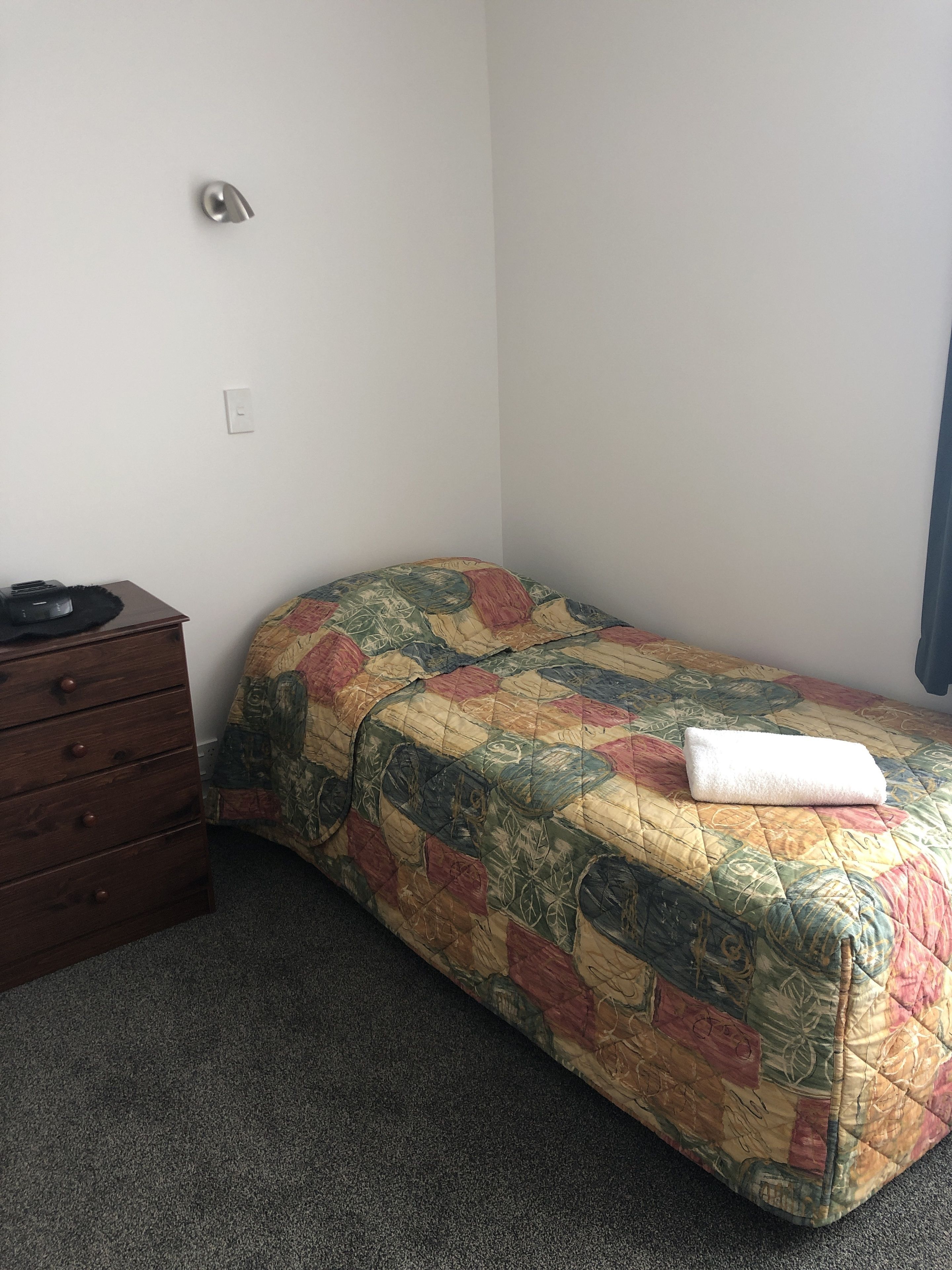 A small, single-bed room with a colorful quilt, a wooden dresser, and a power outlet on the wall.