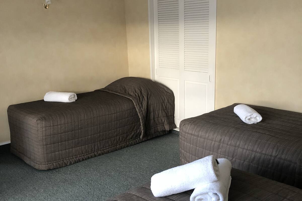 A hotel room with two beds, each with a rolled white towel. The room has beige walls, a white door with louvered shutters, and a carpeted floor.