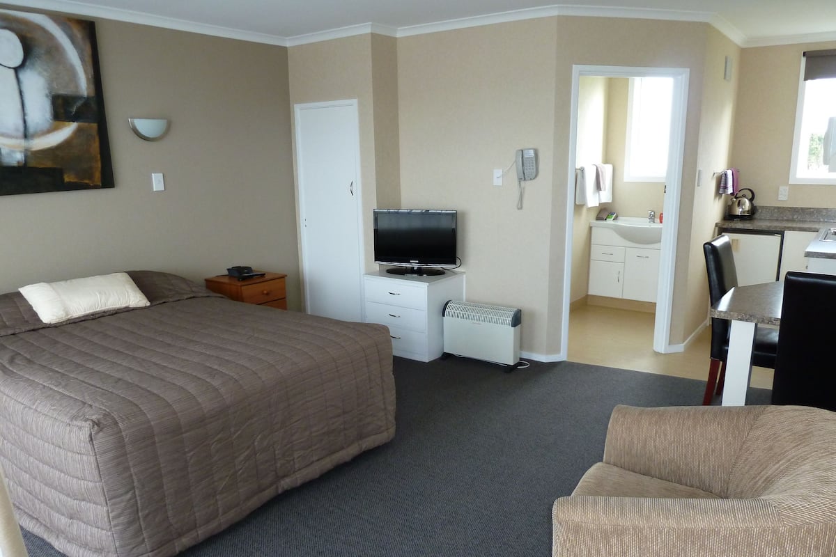 A hotel room with a large bed, a TV, a desk, and a kitchenette.