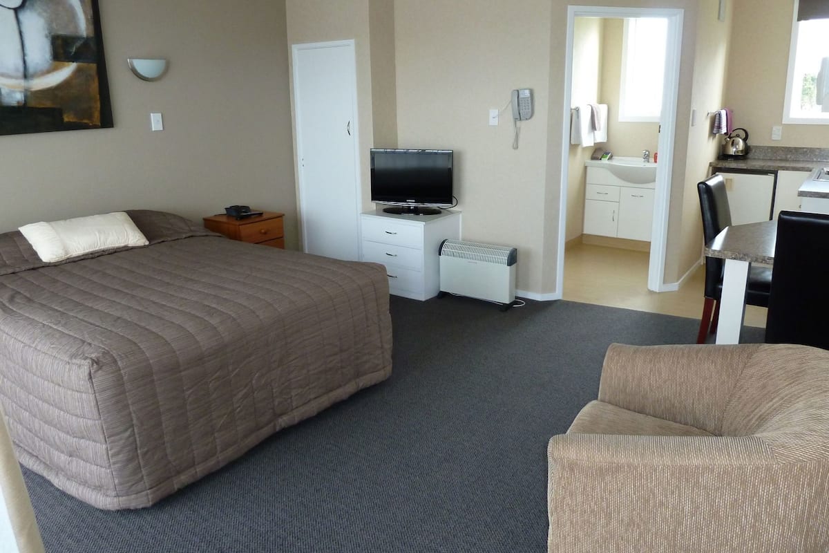A hotel room with a large bed, a TV, a desk with a chair, and a kitchenette.