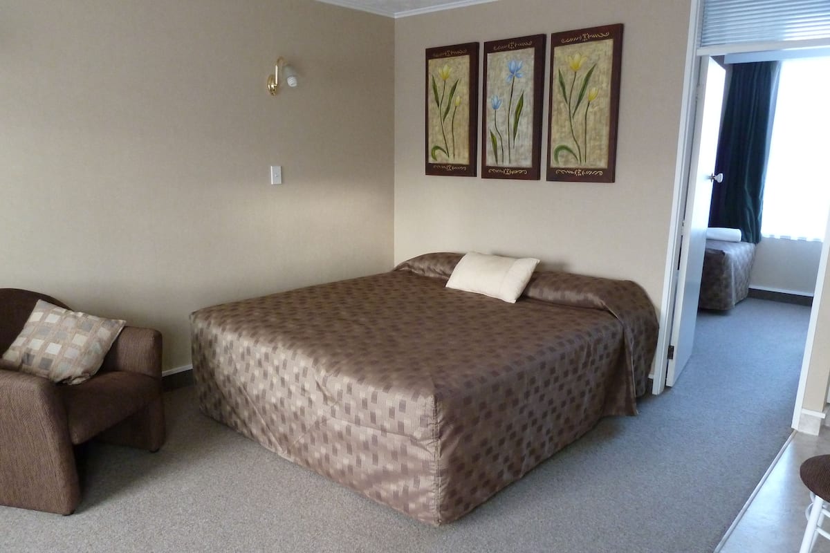 A hotel room with a large bed, a sofa, and a chair. There are three wall-mounted paintings of flowers.