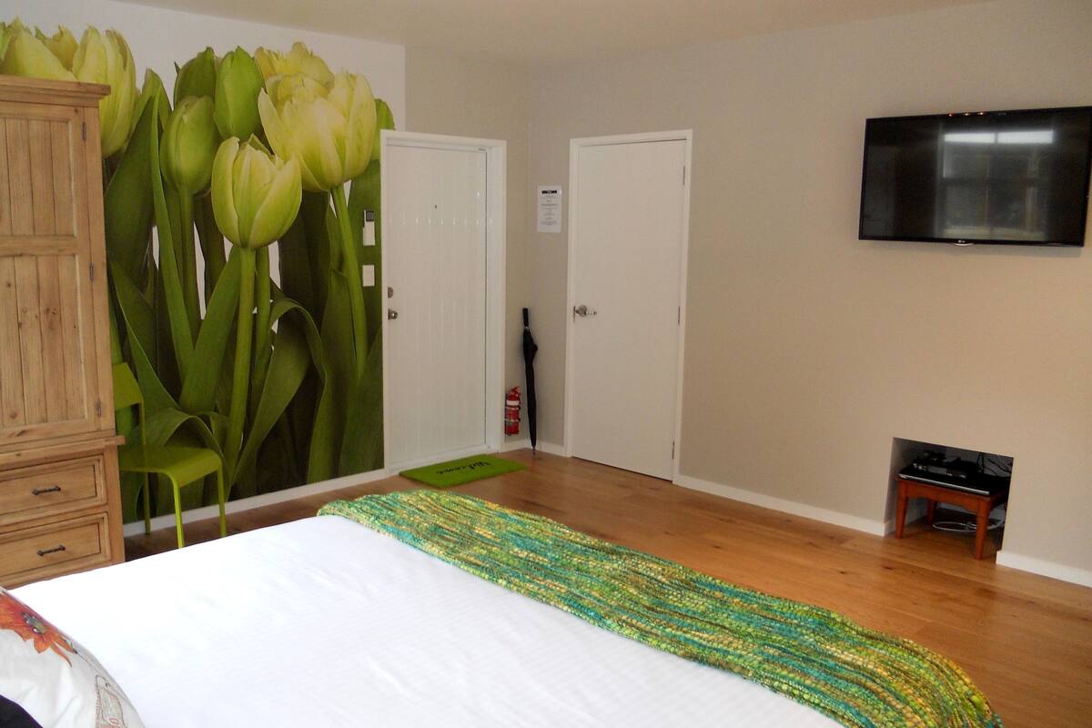 Un dormitorio con cama, armario de madera, un mural de pared verde, televisor y una pequeña mesa de madera.