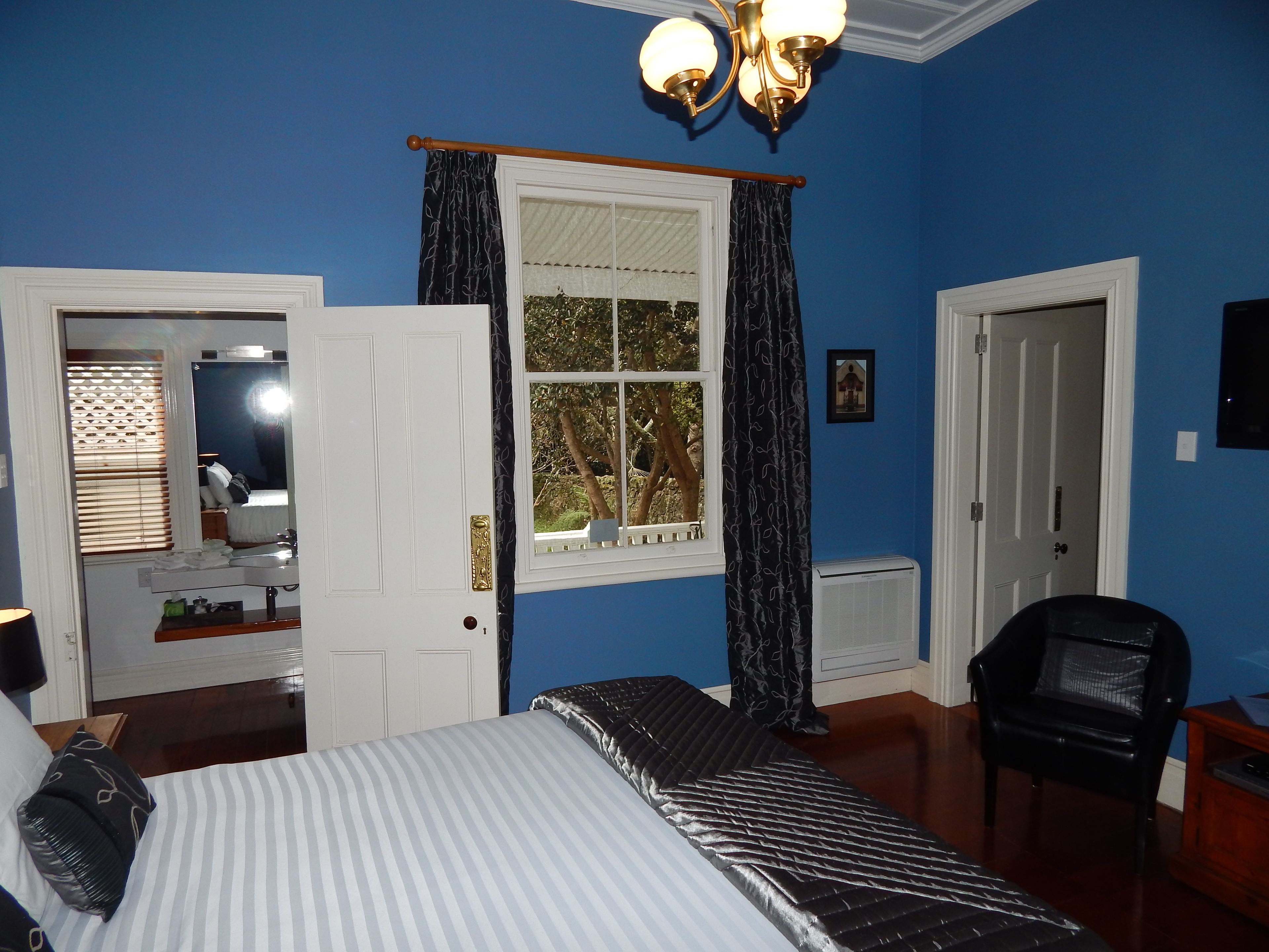 A bedroom with a bed, a chair, a window with curtains, a door, and a mirror.