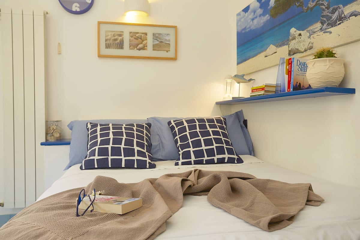 A neatly made bed with a beige blanket, a pair of glasses, and a book on it. There are two pillows with a checkered pattern, a framed picture on the wall, and a shelf with books and a potted plant.