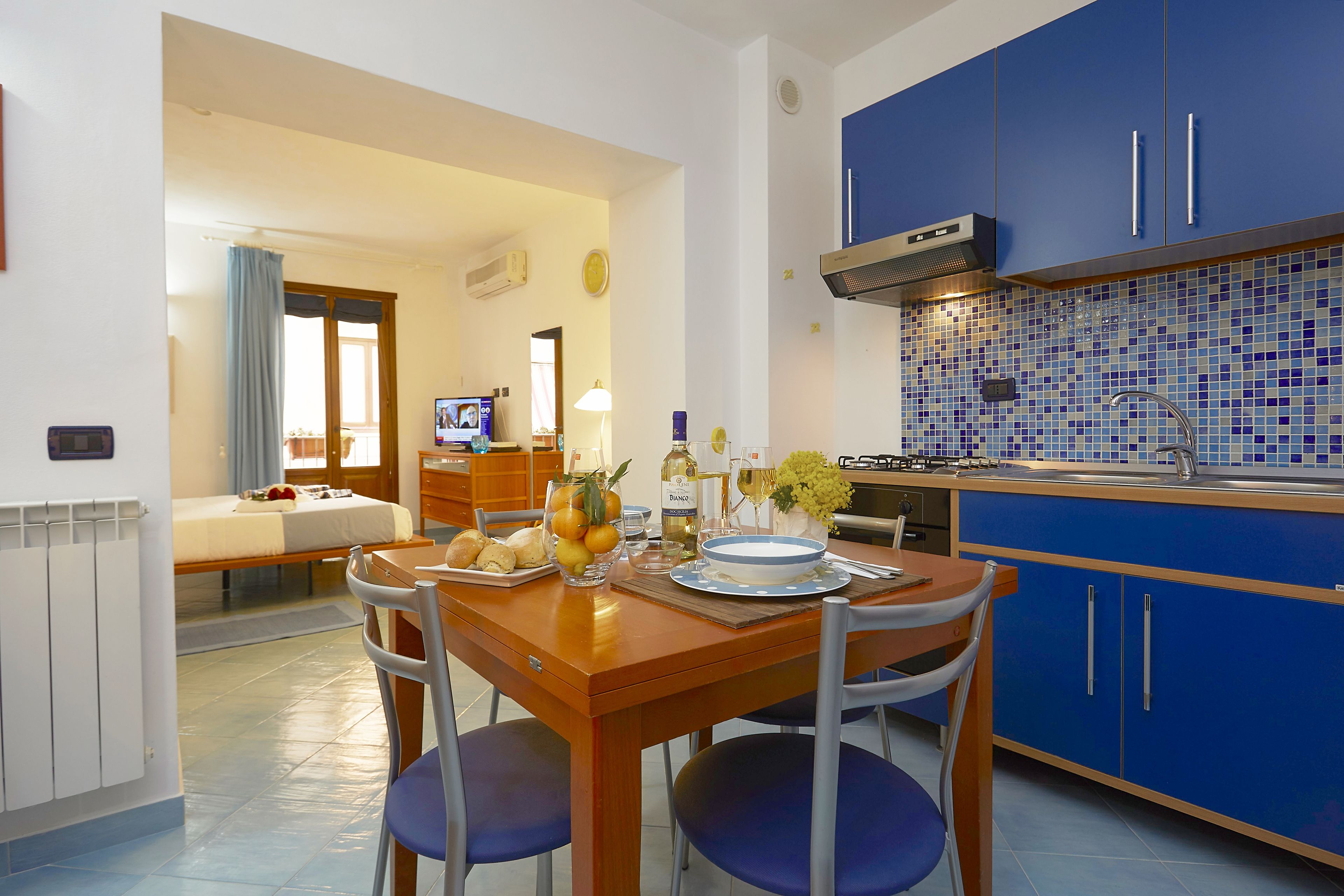 A modern kitchen with blue cabinets, a dining table with chairs, and a TV in the background.