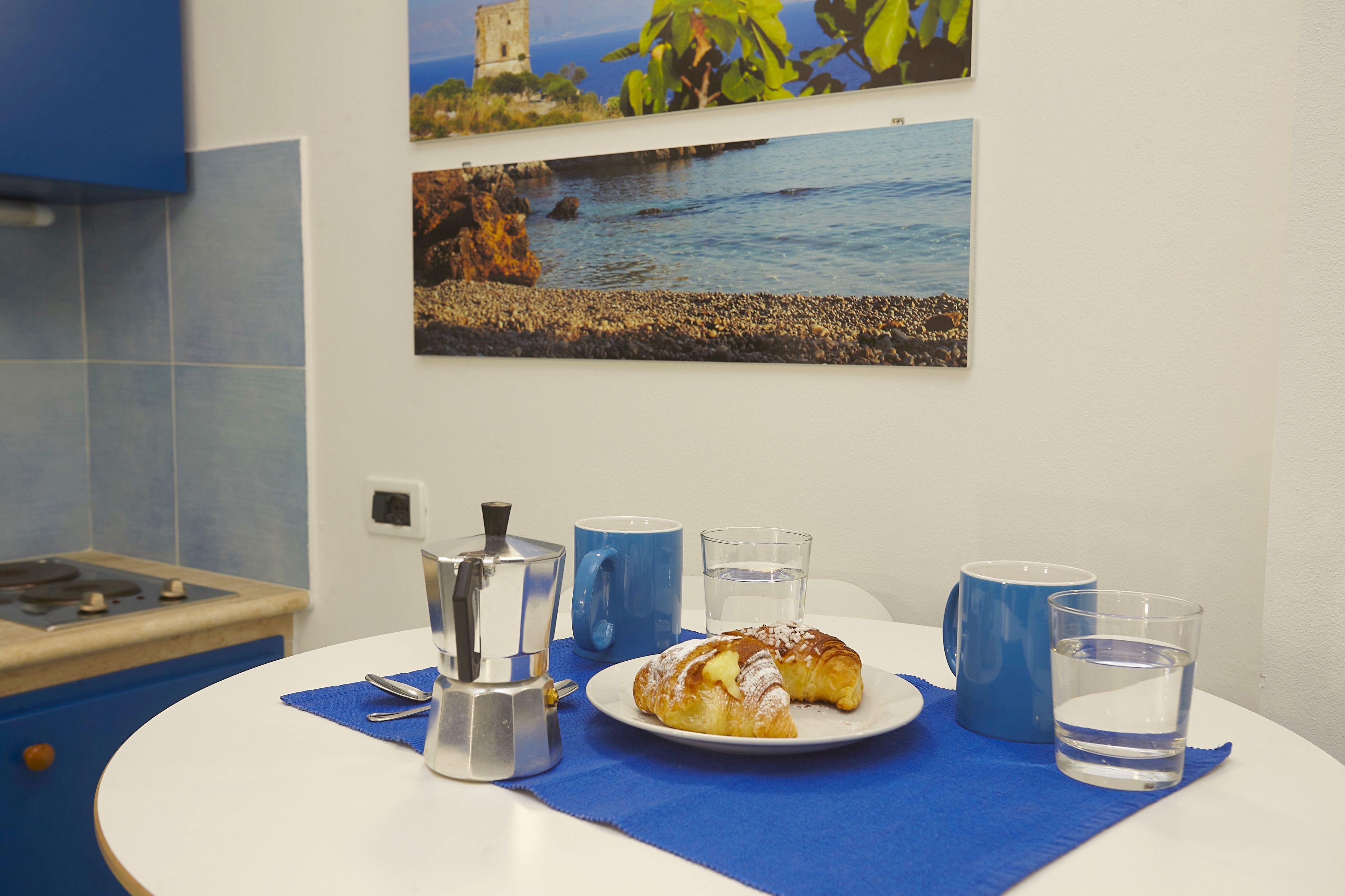 A breakfast setup with a croissant, coffee pot, and two blue mugs on a table with a blue placemat.