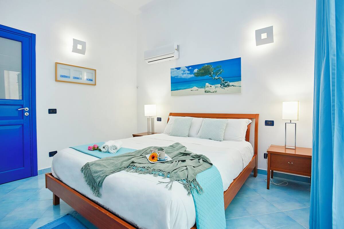 A neatly made bed with a blue blanket, a painting of a beach scene, and a bedside table with a lamp.