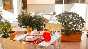 Mesa al aire libre con mantel blanco, platos rojo y blanco y distintos vasos. Hay plantas en macetas alrededor de la mesa, y se ve una silla de madera.