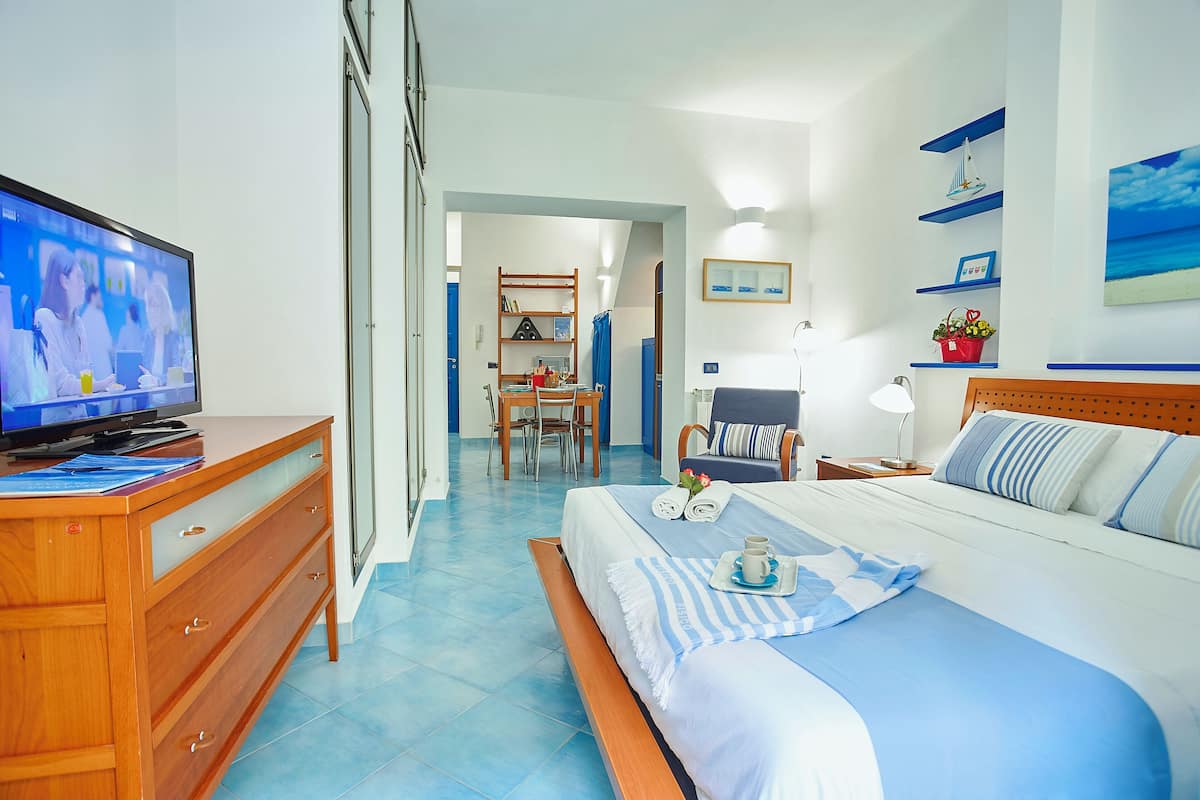 A hotel room with a bed, a desk with a television, a chair, a bedside table with a lamp, and a wall with shelves and a picture.