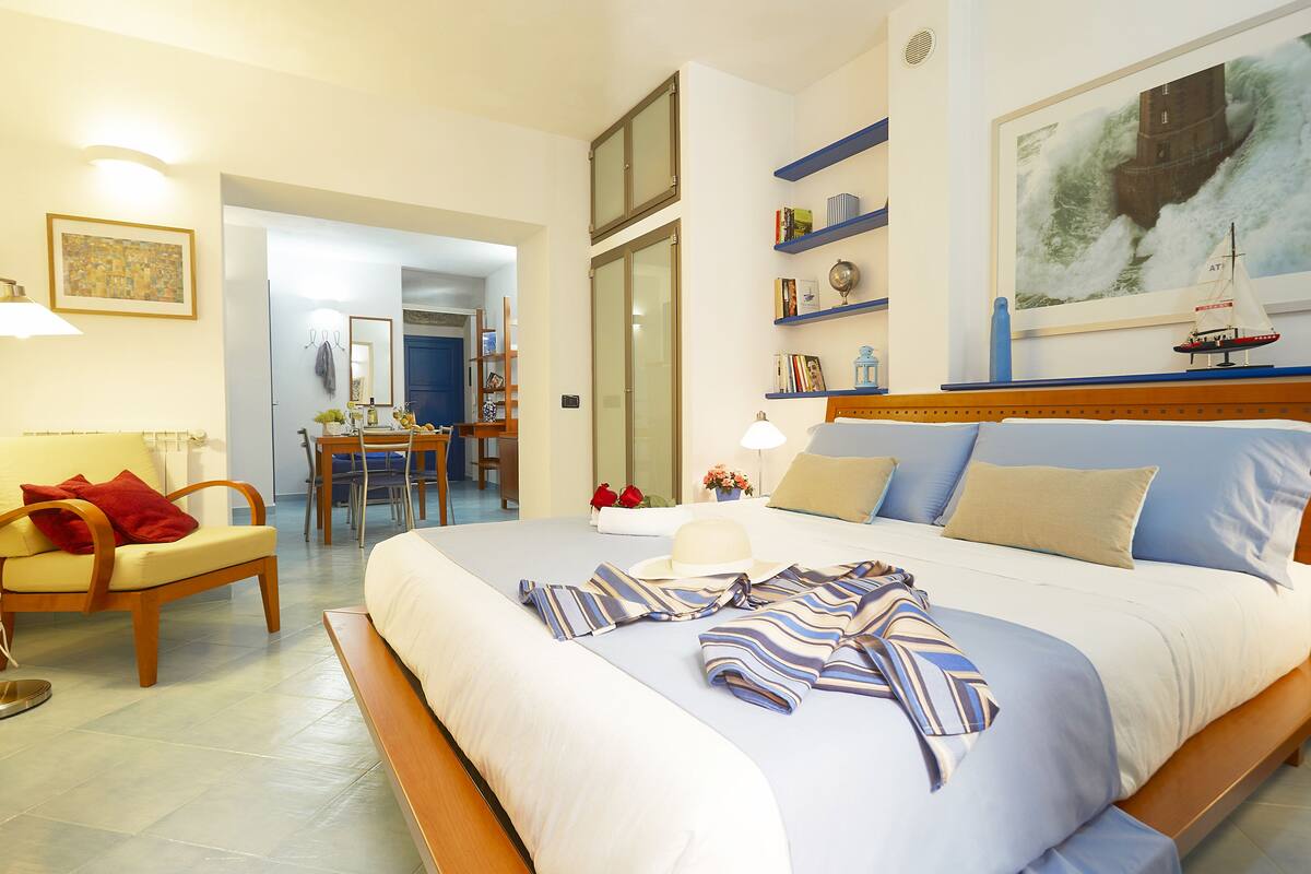A hotel room with a large bed, a desk, and a chair. There is a painting on the wall and a window with a view of the sea.