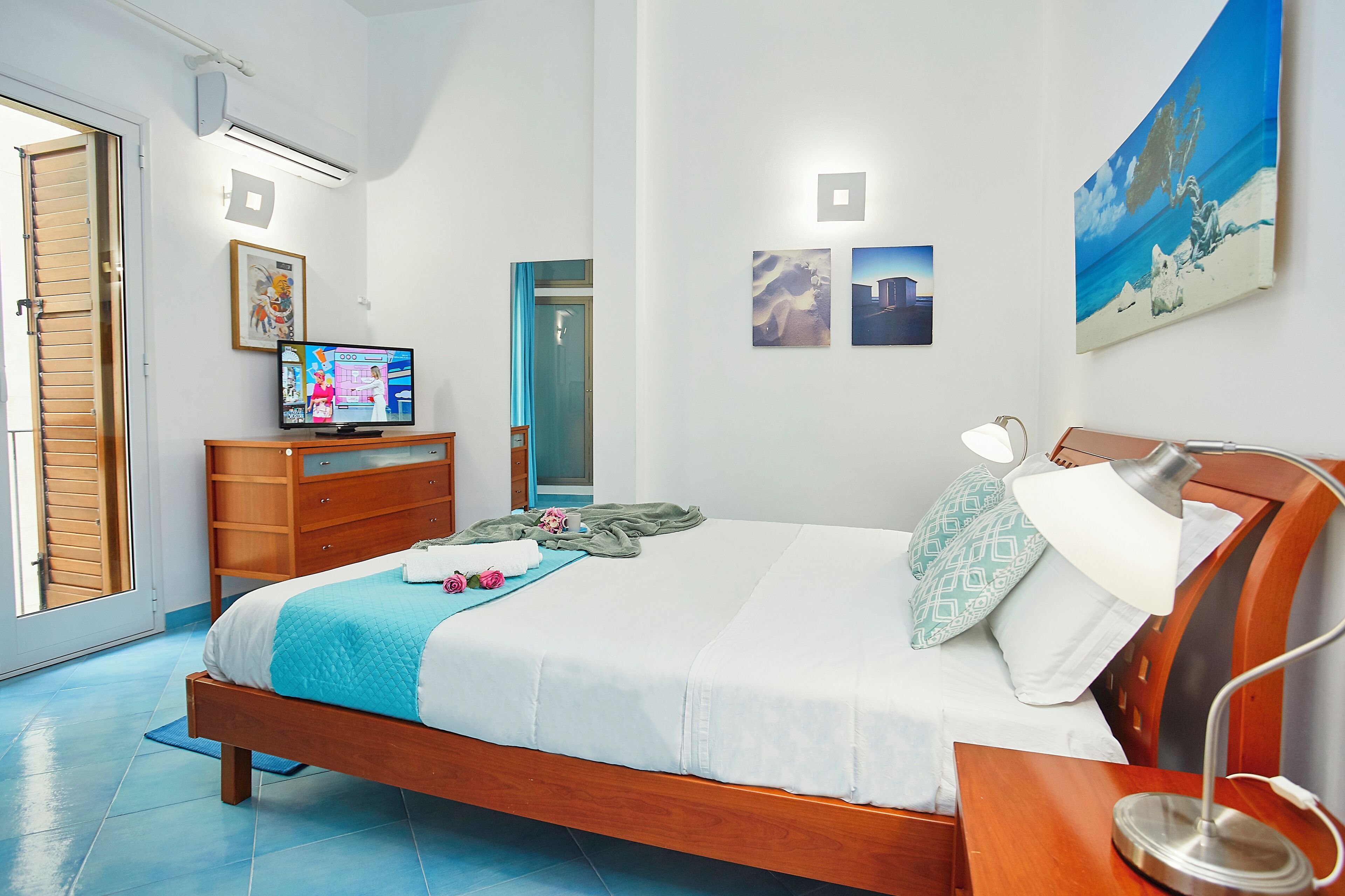 A bedroom with a wooden bed, a nightstand, a desk with a lamp, a television on a dresser, and artwork on the walls.