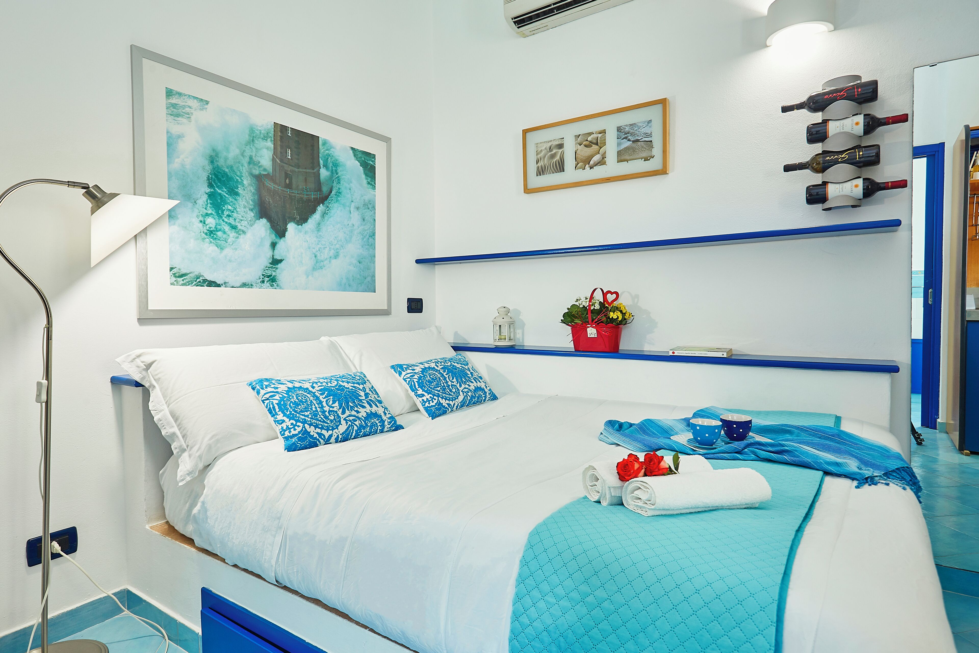 A modern hotel room with a large bed, a framed picture of a lighthouse, a wine rack, and a small plant on the nightstand.