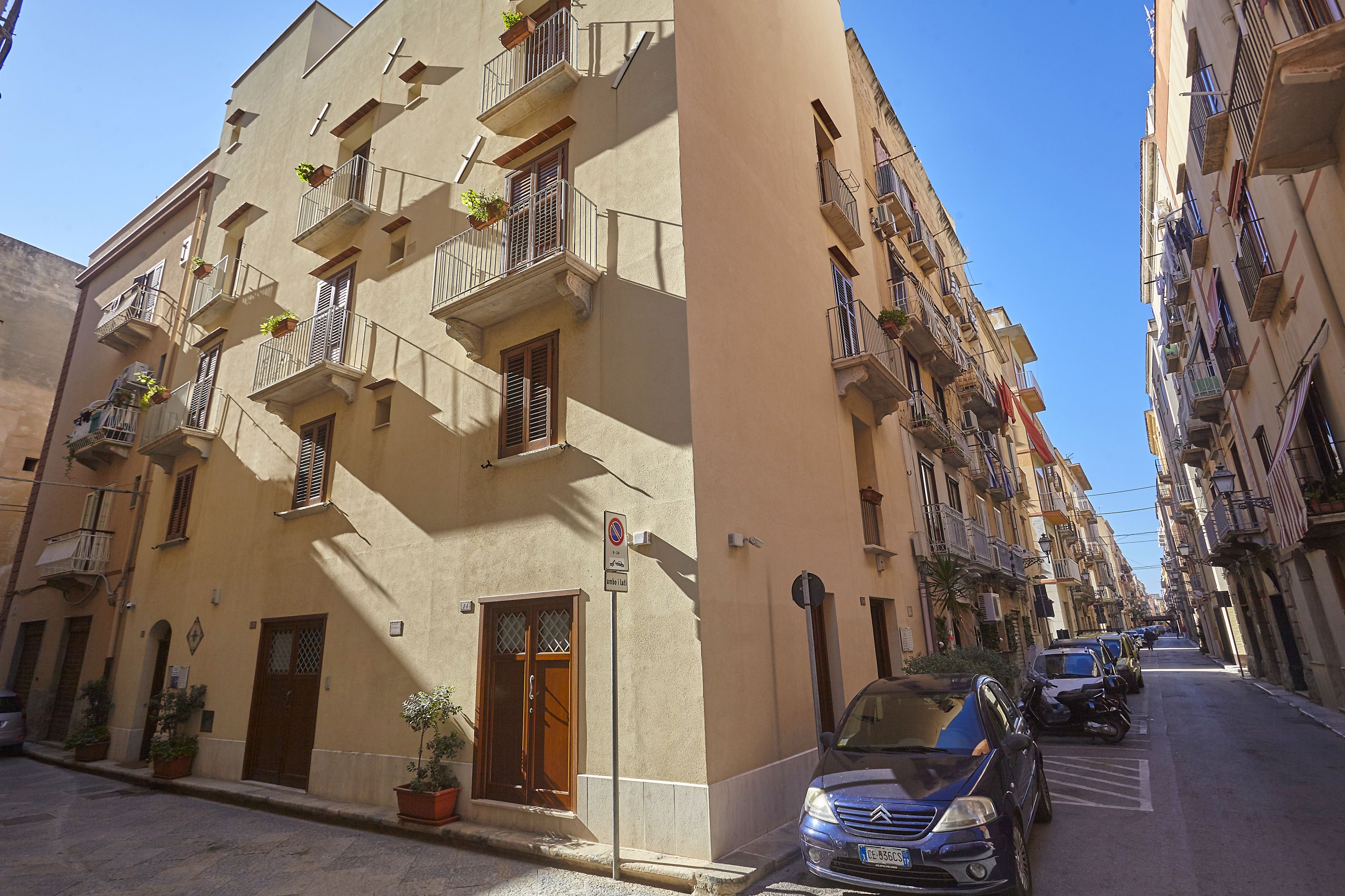 A narrow street with parked cars and a building with multiple balconies, some with plants.