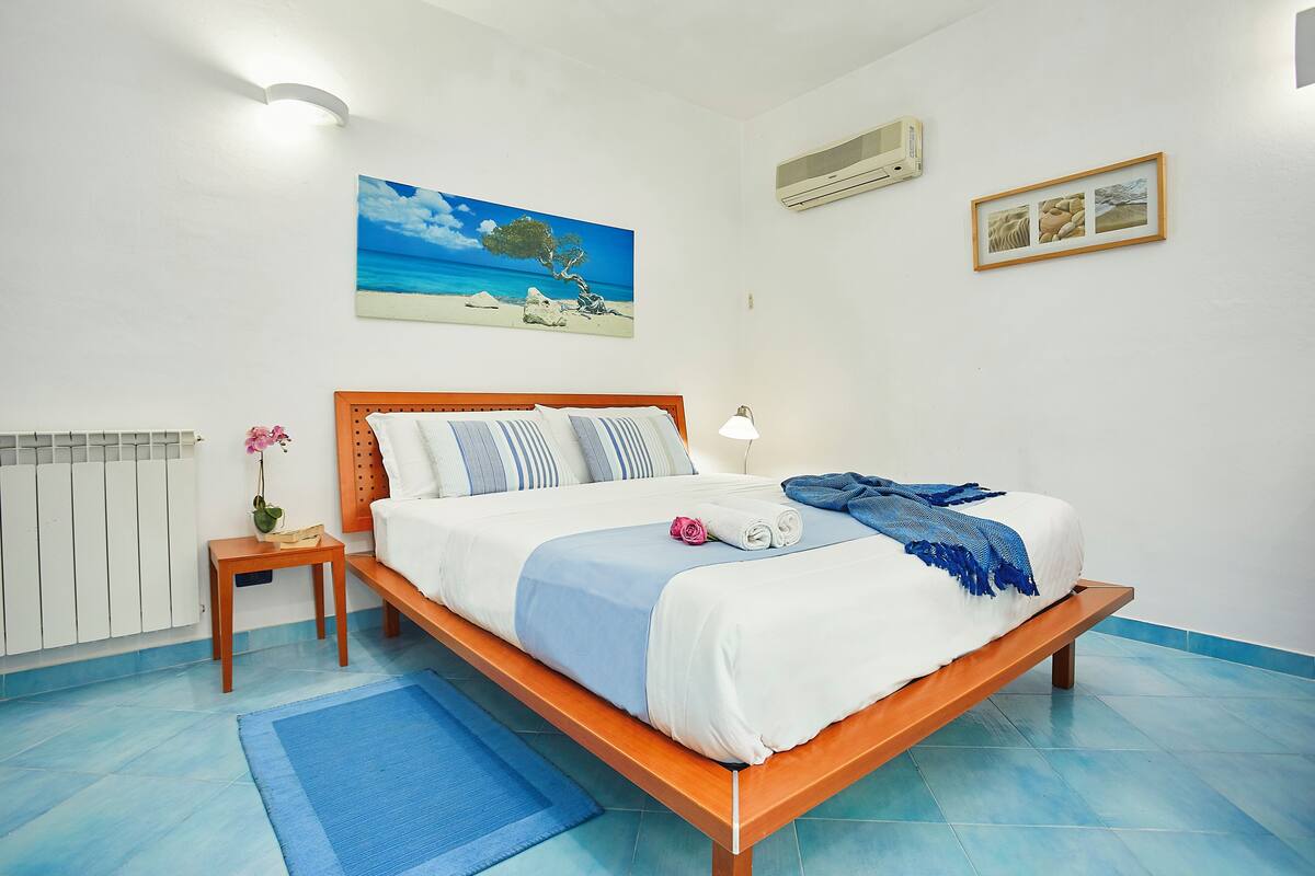A neatly made bed with a wooden frame, a blue and white bedspread, and a blue throw blanket. There is a small bedside table with a lamp and a vase of flowers. A painting of a beach scene hangs on the wall above the bed.