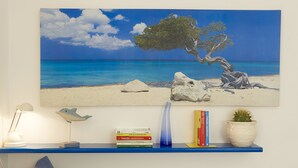 Una pintura que muestra una escena de playa con un árbol, una lámpara, libros y un cactus sobre una estantería.