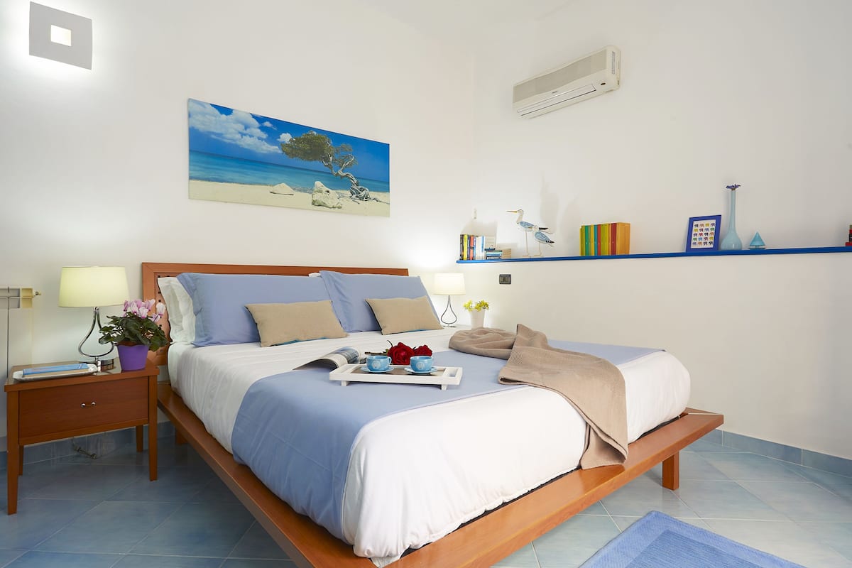 A neatly made bed with a blue and white bedspread, a bedside table with a lamp, a nightstand, a wall-mounted shelf with books and decorative items, and a painting of a beach scene.