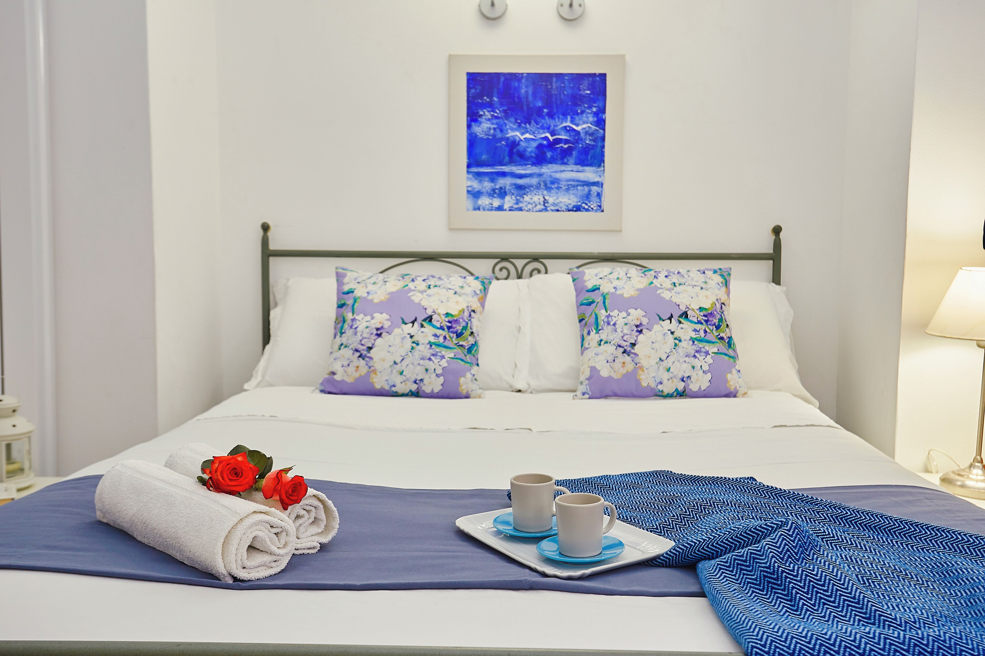 A neatly made bed with floral pillows, a blue blanket, and a tray with two mugs and towels.