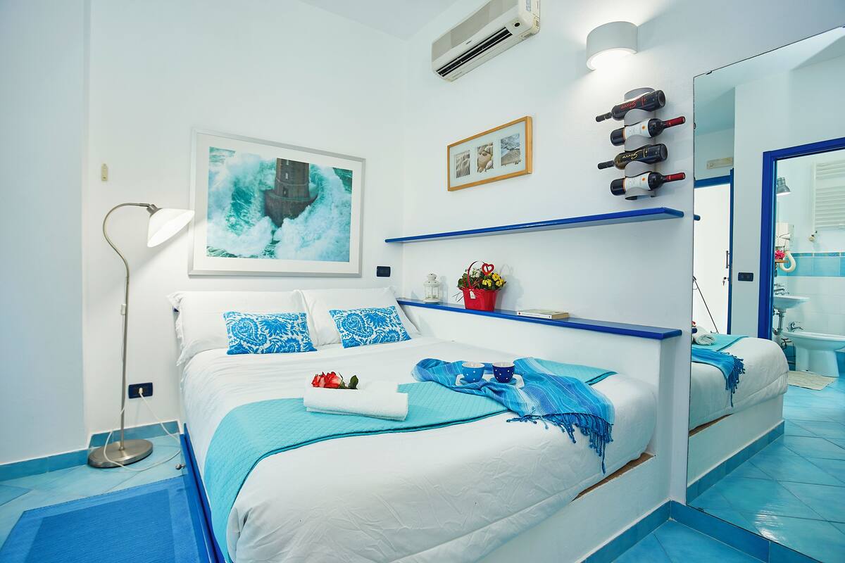 A hotel room with a bed, a bedside lamp, a framed picture, a small potted plant, and a mirror.