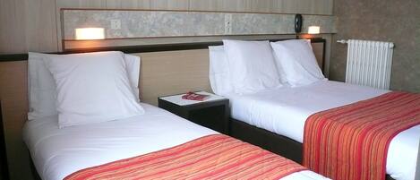 A hotel room with two single beds, each with white linens and a red-striped blanket. There is a nightstand between the beds with a book and a lamp. The room has a wooden headboard and a wall-mounted light fixture.