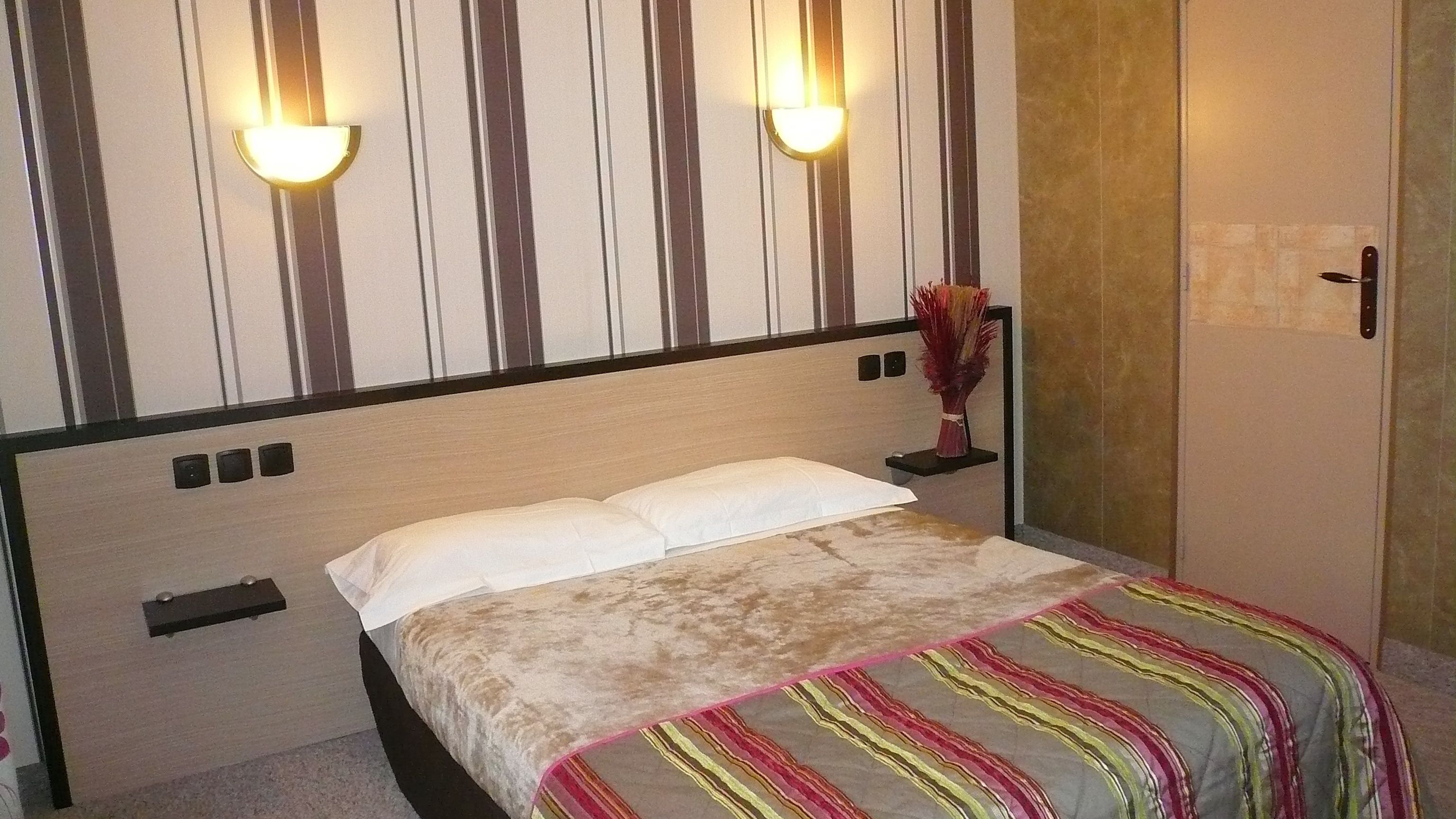 Een slaapkamer met een bed, twee wandlampen, een nachtkastje, een vaas en een deur.