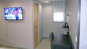 A small room with a TV mounted on the wall, a door, a window with blinds, a table, and a chair. - Hôtel L'Européen (Tarbes)