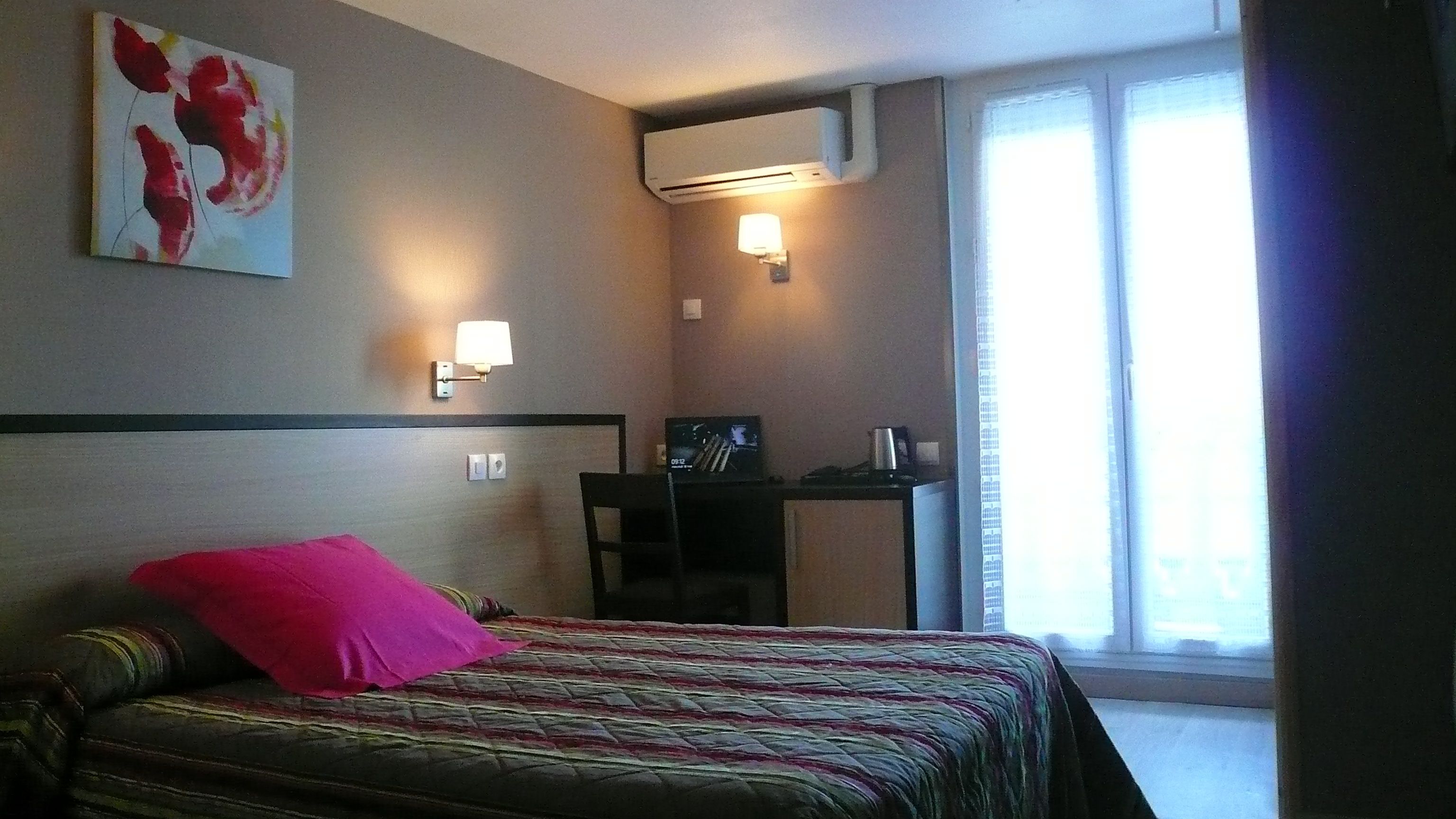 Una habitación de hotel con cama, escritorio, silla, equipo de aire acondicionado y ventana con cortinas.