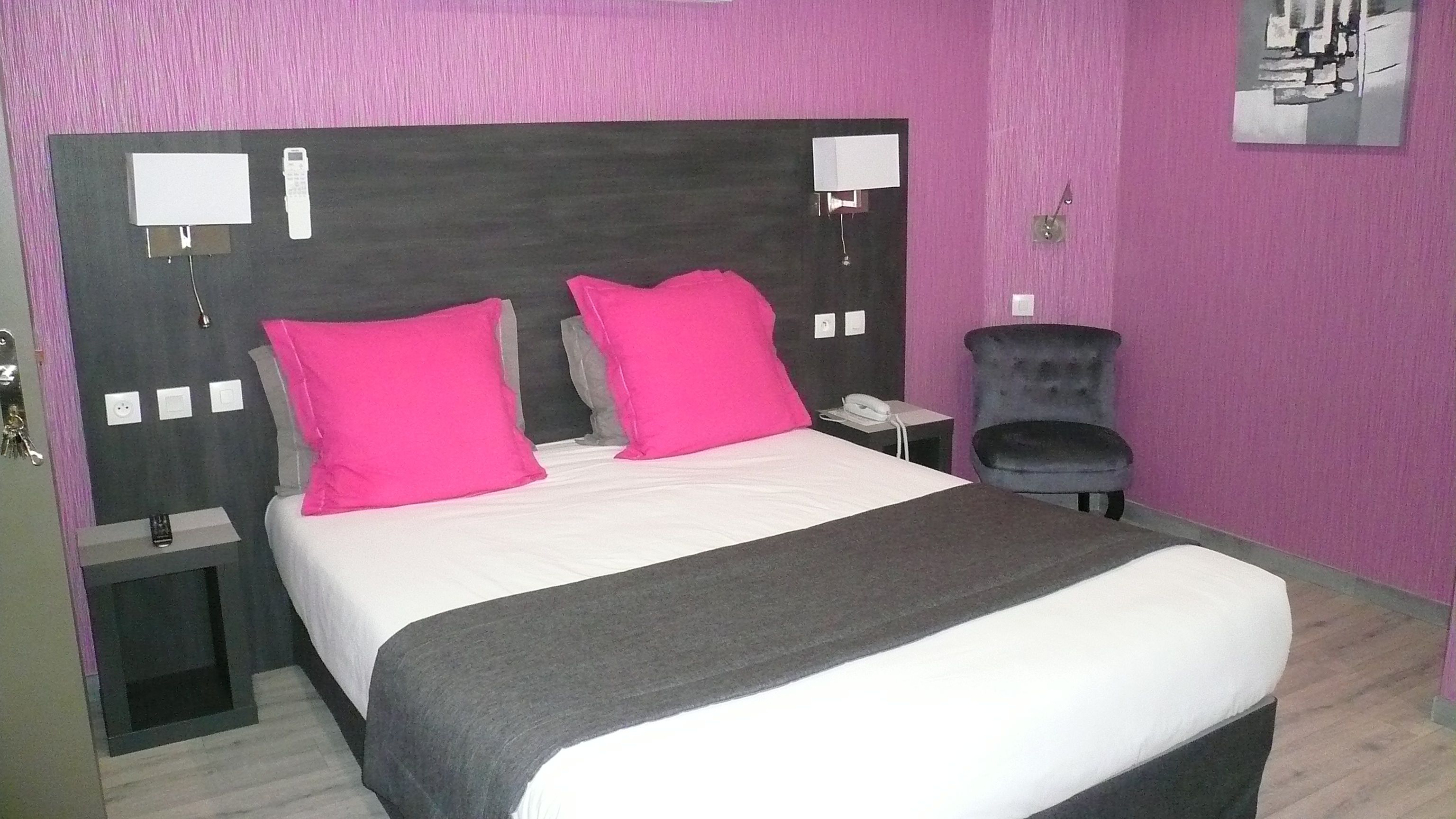 Una habitación de hotel con una cama grande, dos almohadas rosa brillante, un cabecero oscuro, una mesita de noche pequeña con un teléfono y una silla.