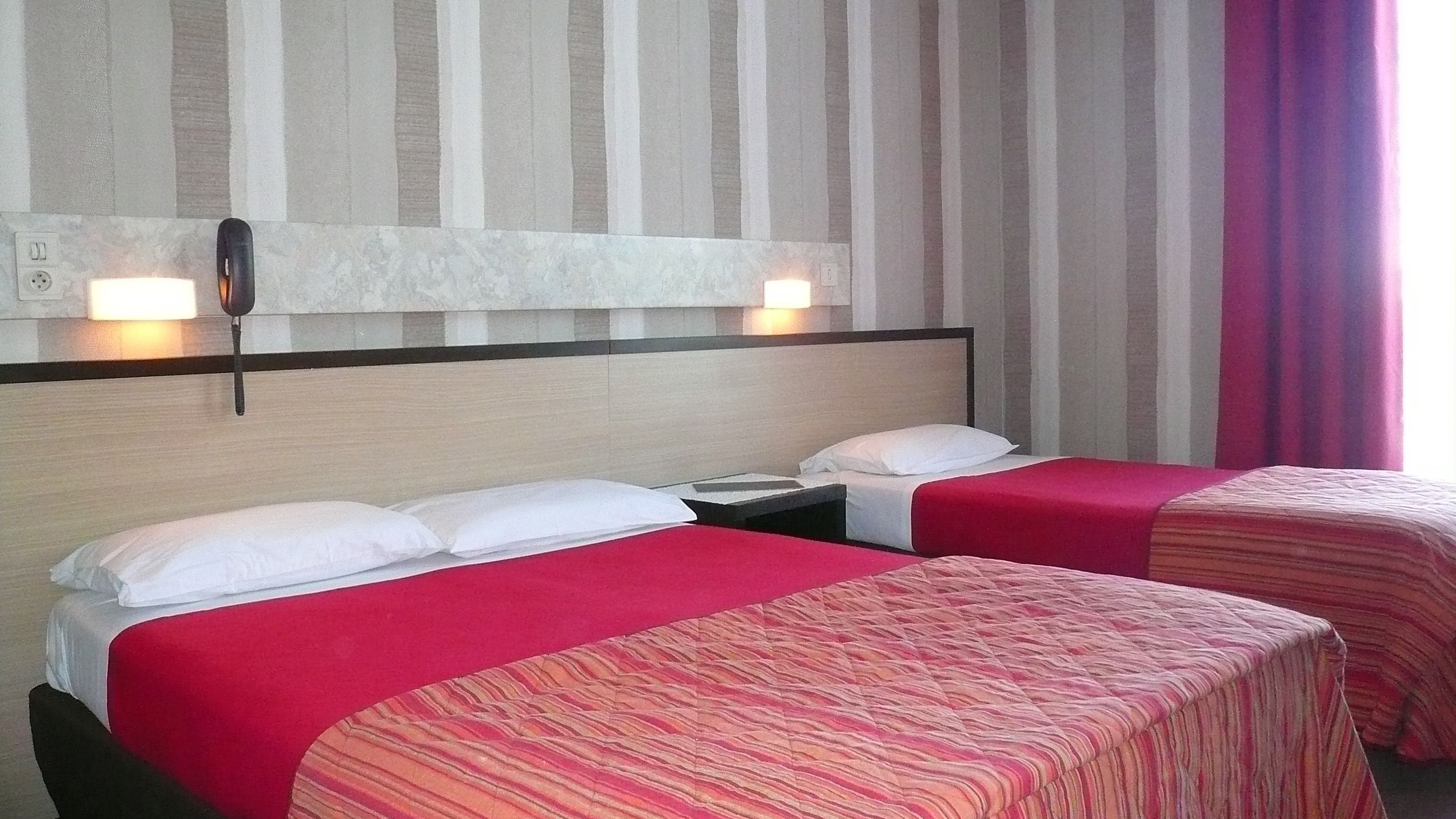 Habitación de hotel con dos camas, cada una con colcha roja y almohadas blancas. Hay un teléfono montado en la pared y una lámpara sobre las camas.