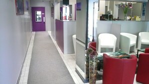 A hallway with a purple door, white chairs, and a reception desk with a vase of flowers. - Hôtel L'Européen (Tarbes)