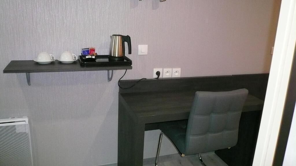 A small kitchenette with a dark countertop, a chair, and a shelf with cups and a kettle.