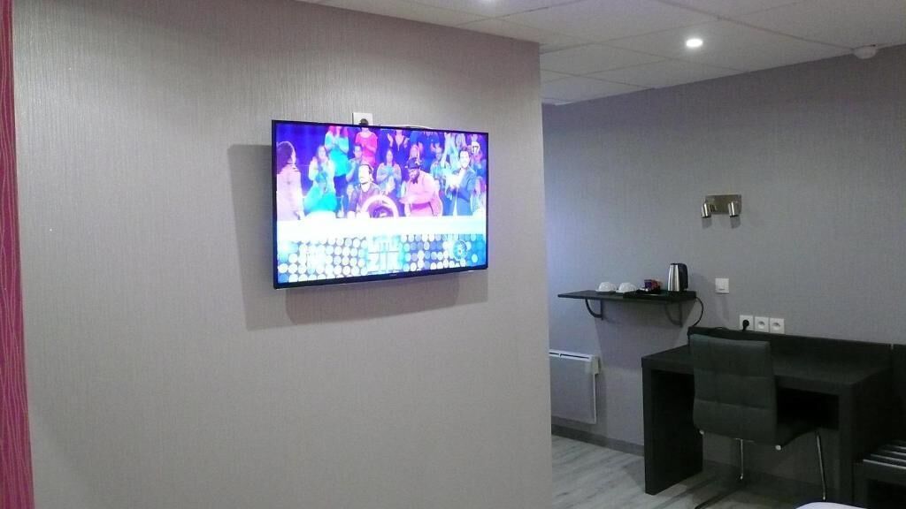 Una habitación con una televisión de pantalla plana montada en la pared mostrando un evento deportivo, un escritorio con una silla y un estante con objetos.