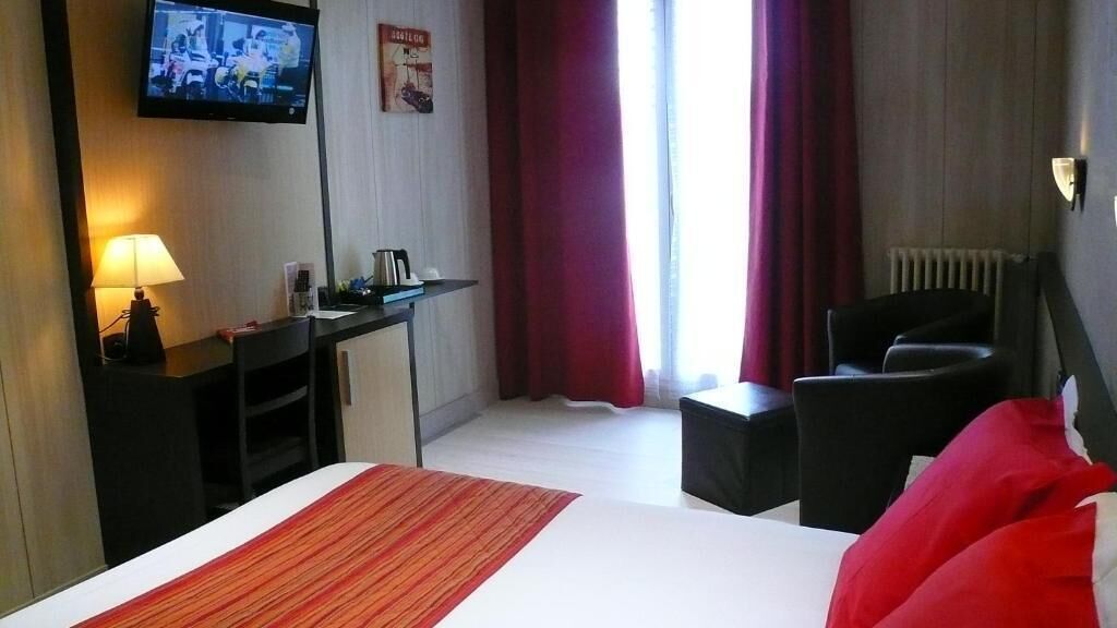 Habitación de hotel con cama, escritorio con lámpara, silla, televisor y ventana con cortinas.