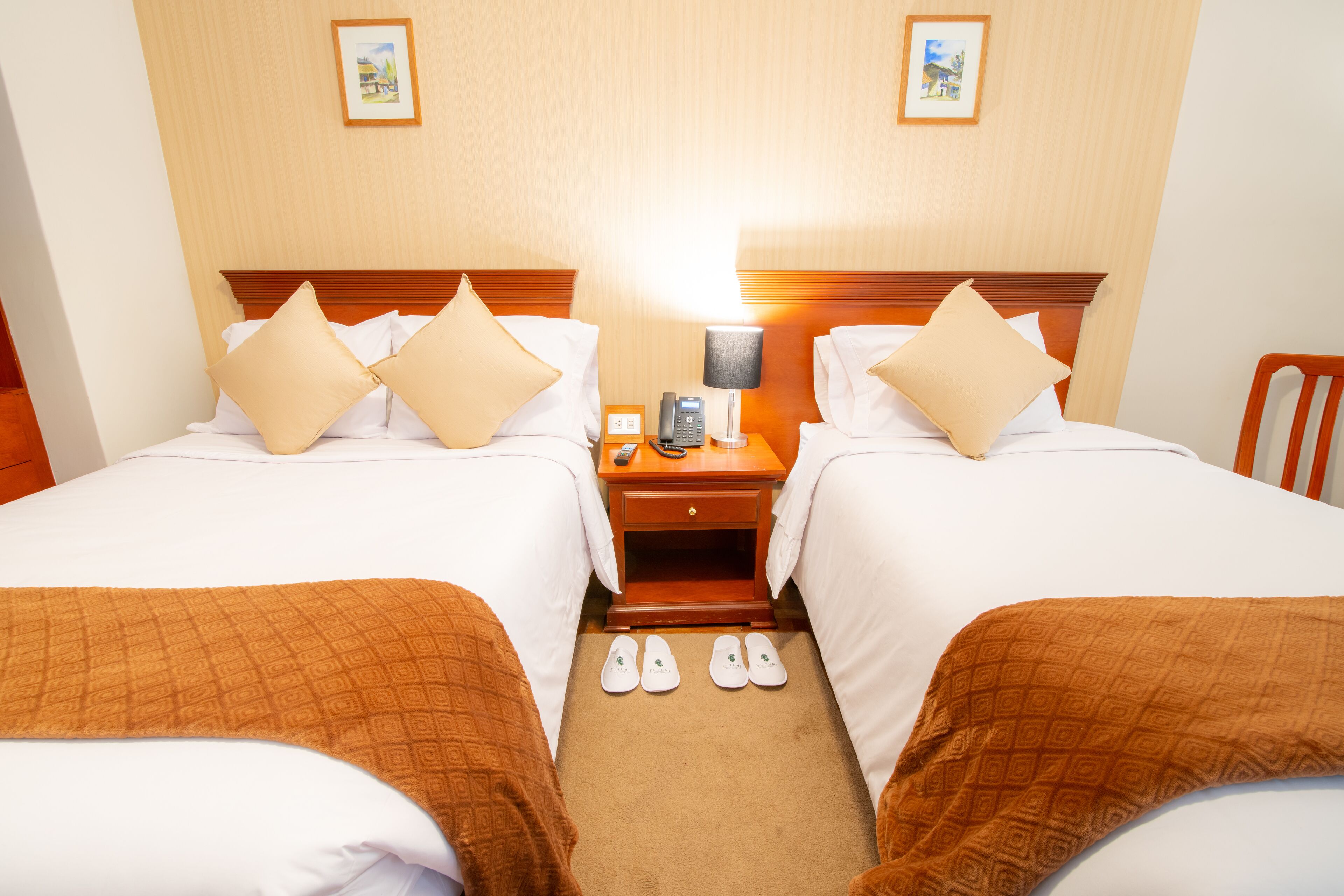 Quarto de hotel com duas camas, uma mesa de cabeceira com abajur e um par de chinelos no chão.