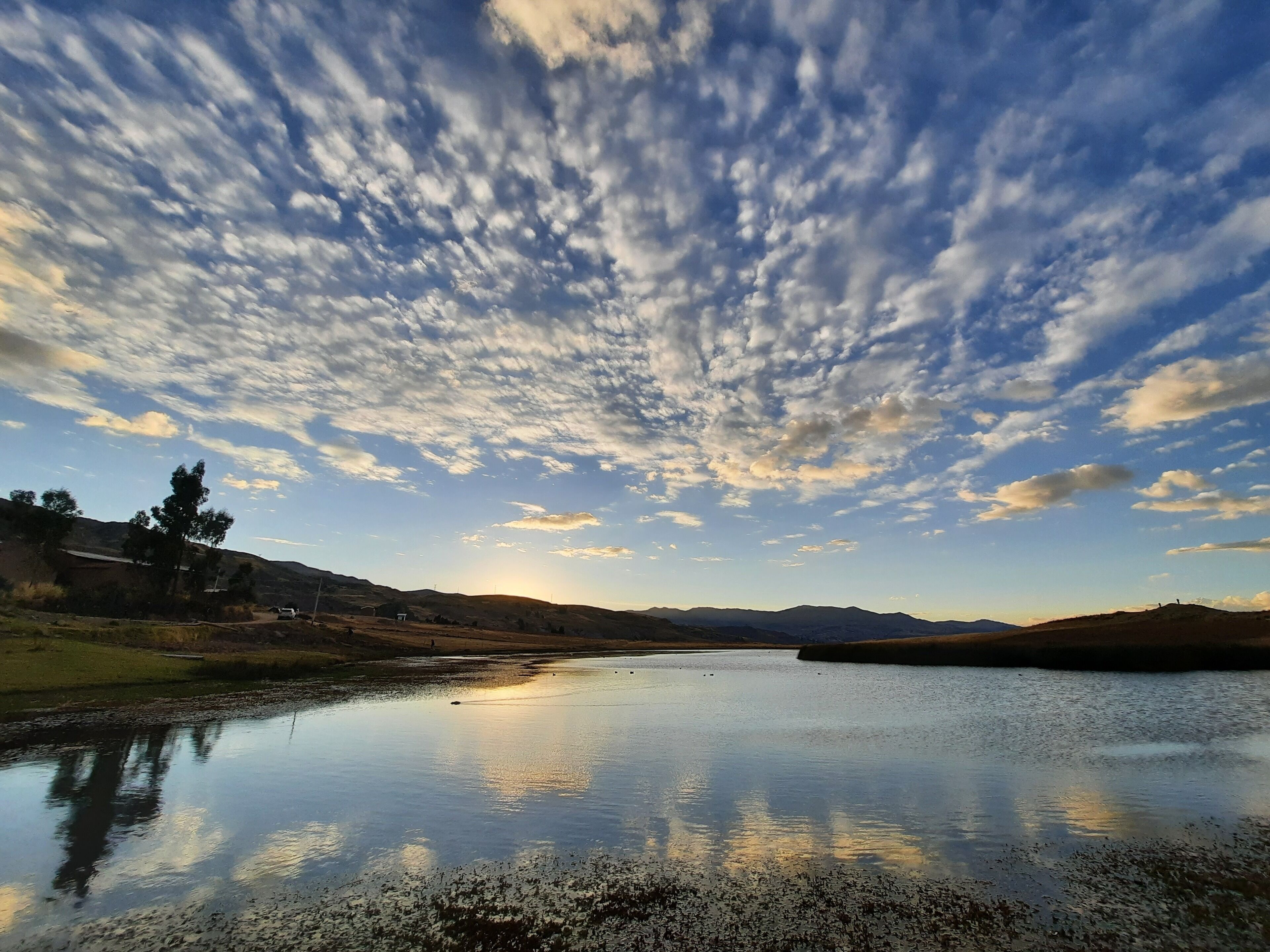 Un lago sereno que refleja el cielo con nubes dispersas, rodeado de colinas cubiertas de hierba y algunos árboles.
