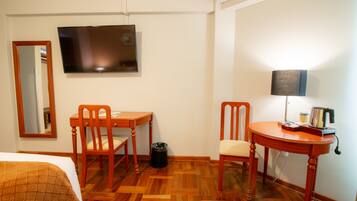 Une chambre d’hôtel avec un lit, un bureau en bois, deux chaises, une télévision, une lampe et une poubelle.