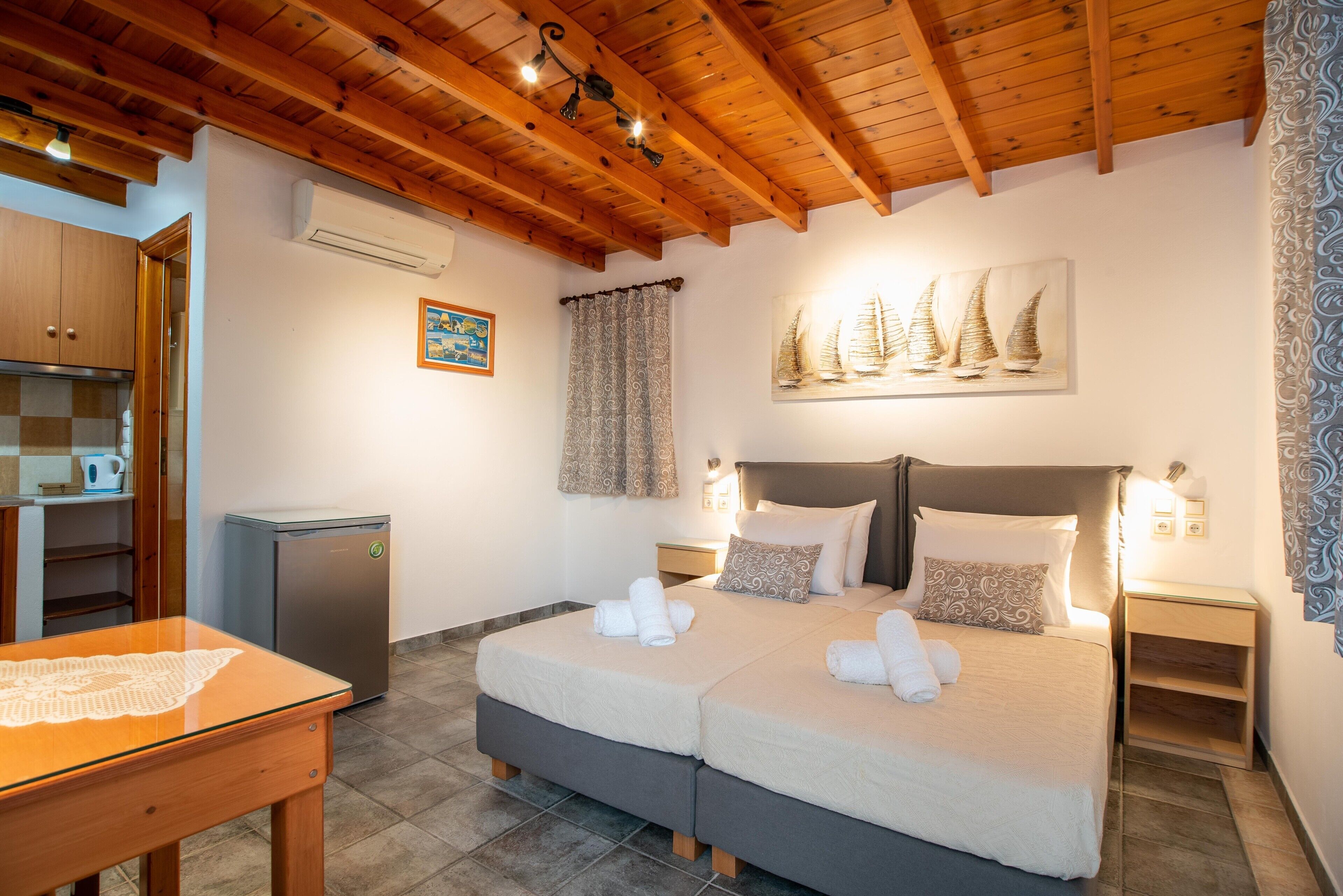 A double bed with white linens and pillows, a small table with a lace tablecloth, a refrigerator, a wooden ceiling, and a painting of sailboats on the wall.