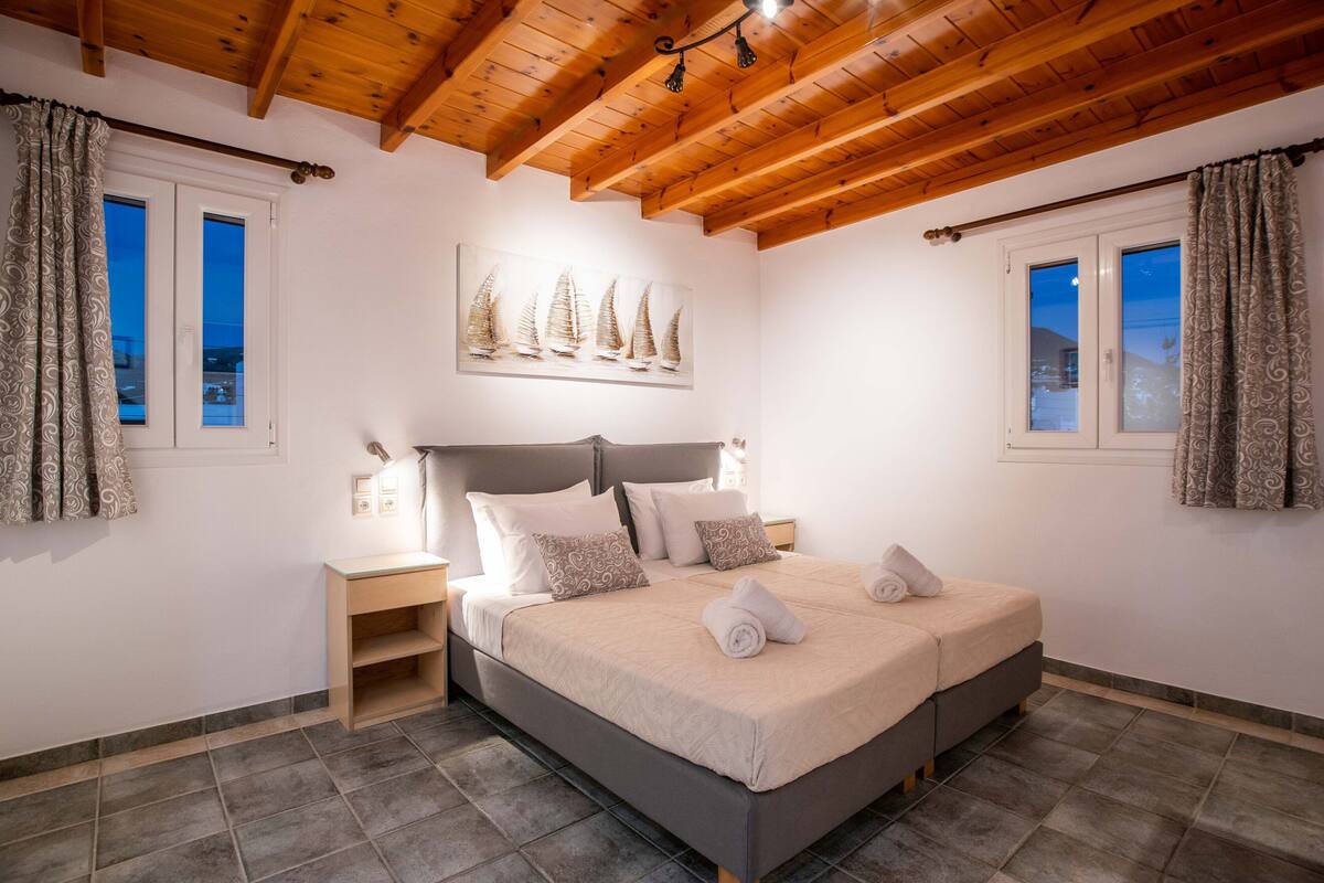 A bedroom with a bed, wooden ceiling beams, a nightstand, a wall-mounted painting, and a window with curtains.