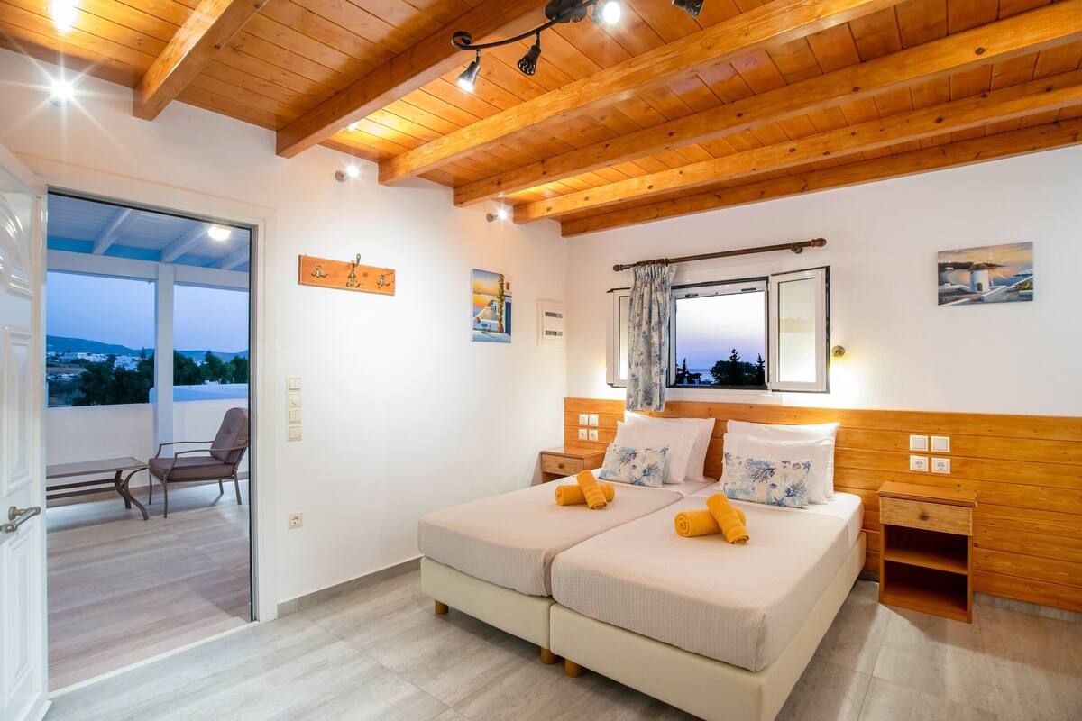 A bedroom with two beds, wooden ceiling beams, a balcony, and a view of the landscape.