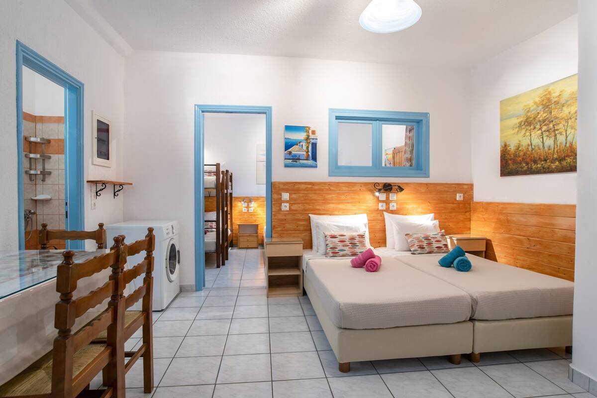 A bedroom with a double bed, wooden headboard, a wooden table, a chair, a washing machine, and a bathroom with a shower and a mirror.