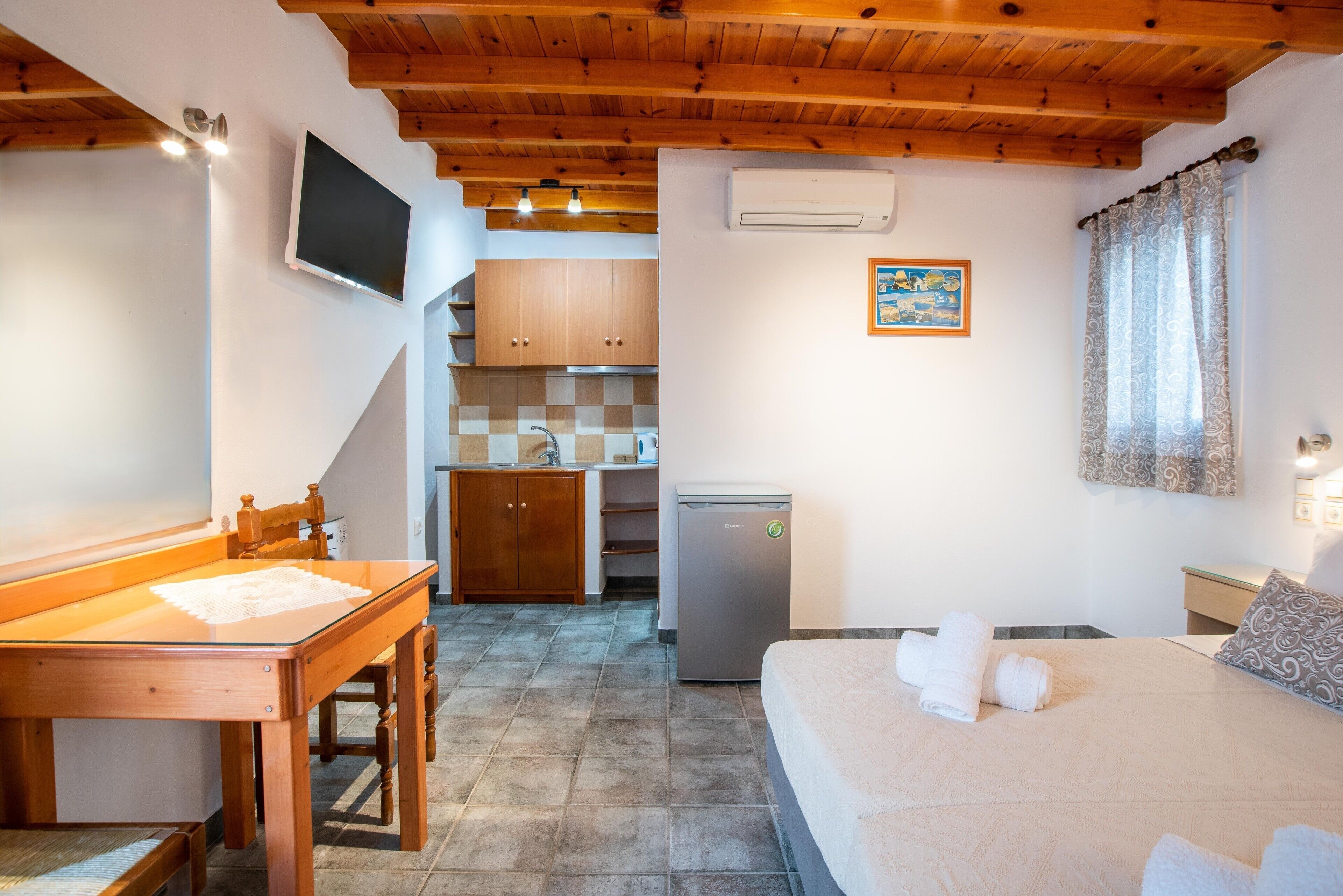 A bedroom with a bed, a desk, a chair, a television, a refrigerator, and a kitchen area in the background.