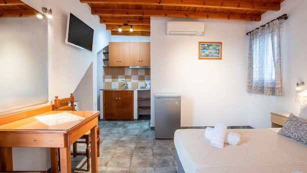 A bedroom with a bed, a desk, a chair, a television, a refrigerator, and a kitchen area in the background. - Kontaratos Studios & Apartments (Paros)